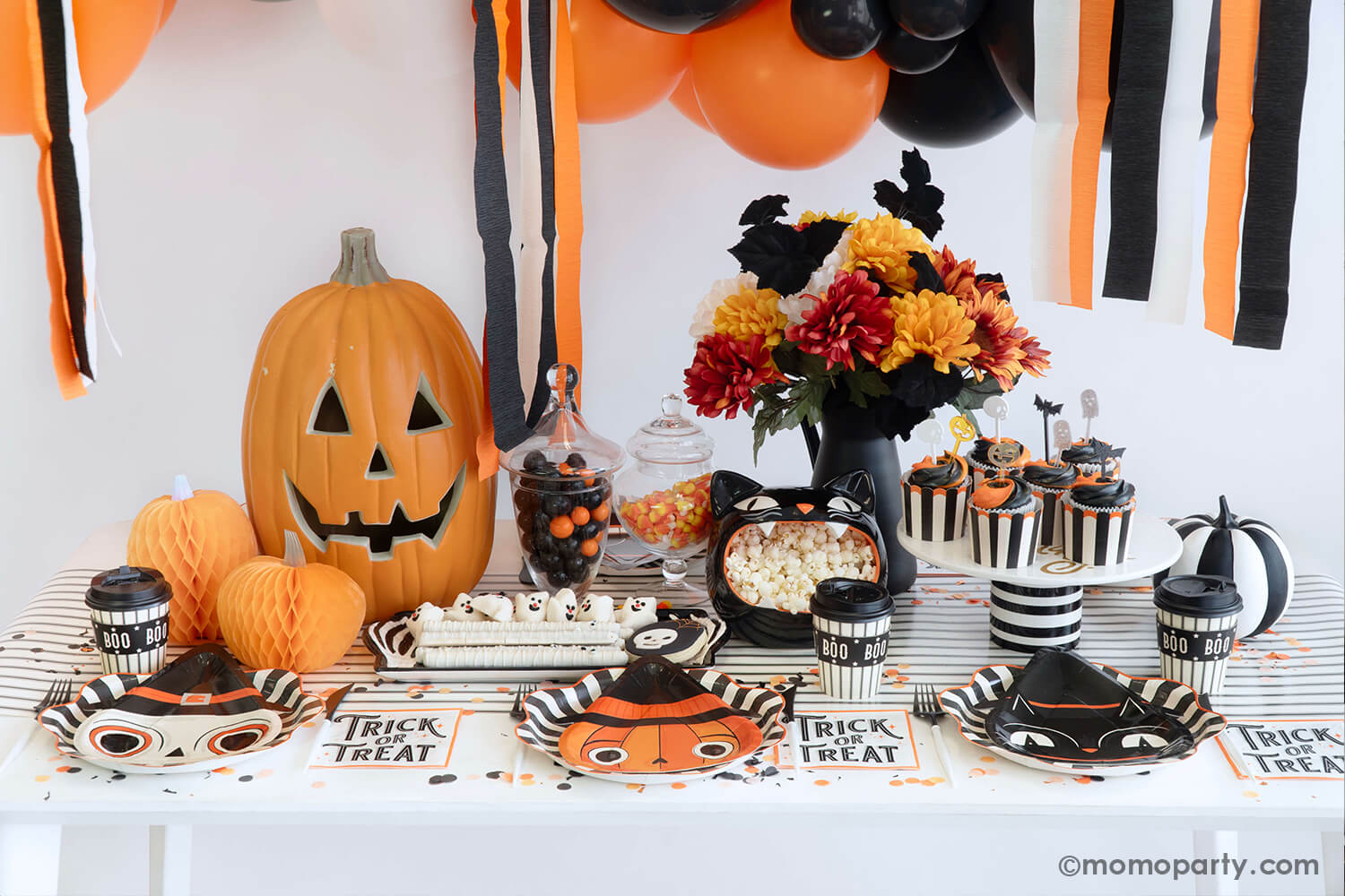 A vintage Halloween party table featuring various party supplies from Momopartys including black striped starburst dinner plates, vintage Halloween character shaped side plates including the skull, the pumpkin and the black cat, Boo! to-go party cups, trick or treat small napkins, black striped food cups, honeycomb pumpkin decorations and monster mash acrylic cupcake toppers. With Halloween treats and fall flower arrangement, this makes a fun and festive kid's friendly Halloween bash in this spooky season!