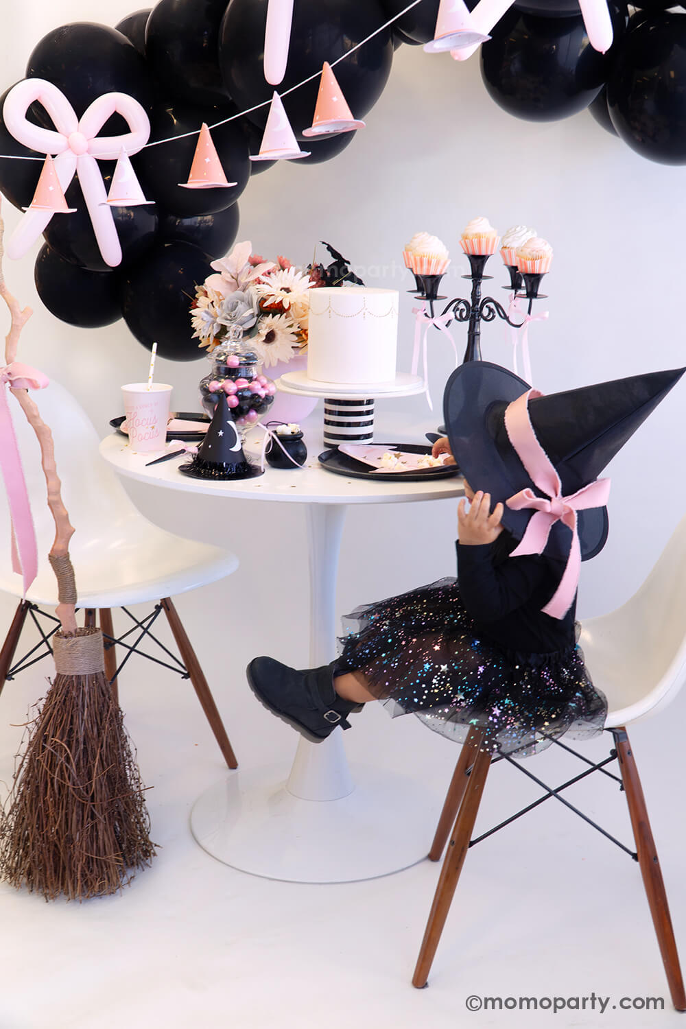 A little girl dressed as Halloween witch with an adorable pink bow on her witch hat setting at a cute witch-inspired Halloween party table featuring various pink Halloween party supplies from Momopartys including pink witch hat shaped plates, Hocus Pocus pink party cups, Hocus Pocus pink small napkins, with a pink floral arrangement and a black candle holder holding pink cupcakes on the table. In the back there's a black balloon garland adorned with pink bow balloons, with a broom adorned with a pink bow. 