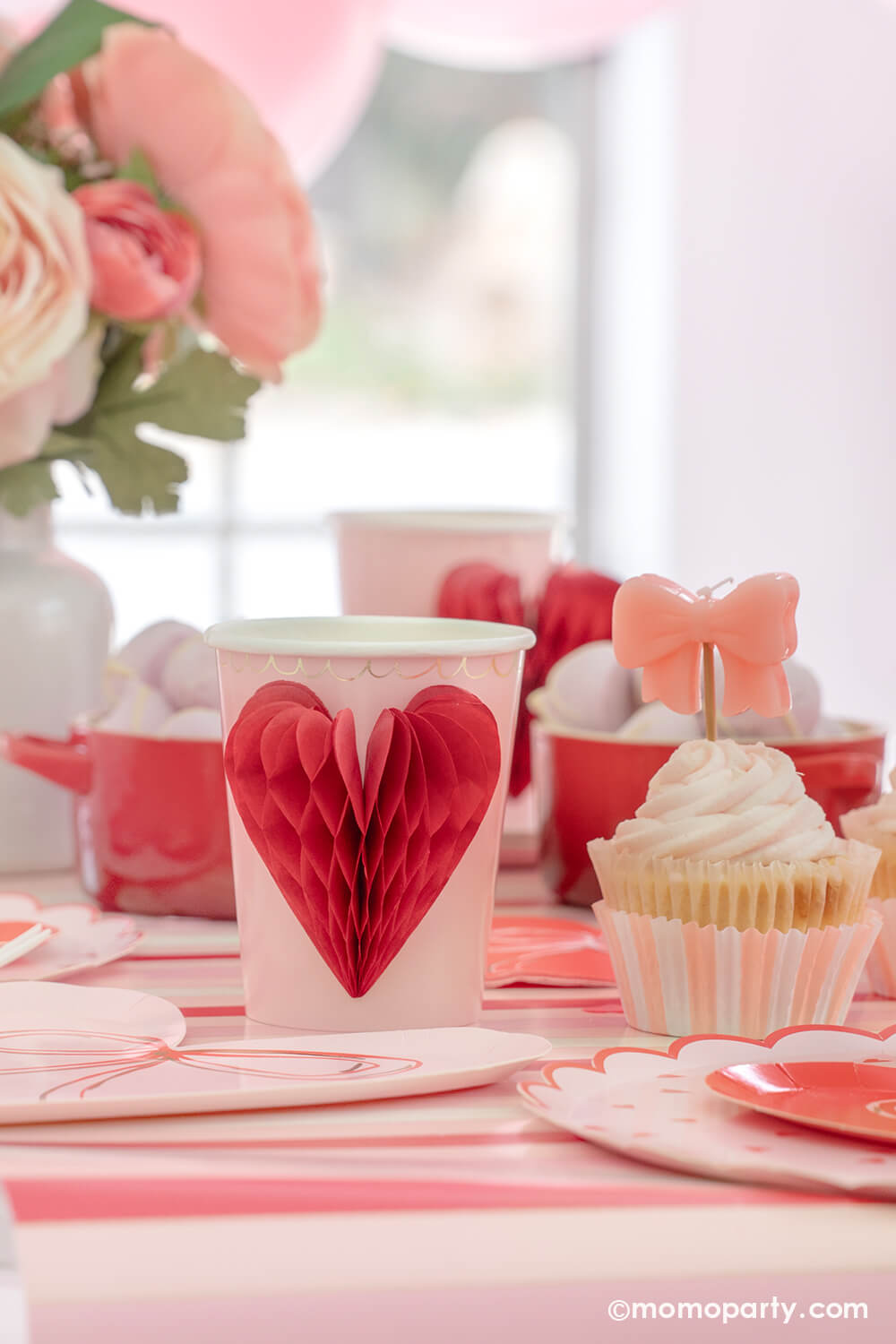 A close-up detail of Momopartys's sweet Pink and Red Bow-Themed Valentine's Day Party table. Highlighting Meri Meri Honeycomb Heart Cups, these cups feature charming 3D honeycomb red hearts on pink paper, adorned with gold foil details. Accompanied by a Pink Bow Candle atop a cupcake and sweets in the red Mini Cocotte, with a flower vase behind them. All elegantly arranged on a red-striped paper runner. Such a cute idea for elevating your Valentine's celebration or Galentine's Day with your besties.
