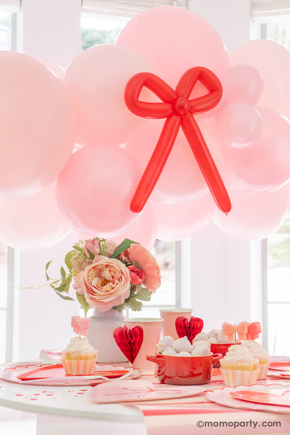 A close up shot of Momopartys's Bow-tiful Valentine's Day Party Box for 8 guests. Featuring a pink balloon garland with red bow balloons on it, heart shaped tableware including plates, napkins and party cups, and a beautiful flower bouquet, this adorable party box in classic Valentine's colors of red and pink is perfect for your Valentine's Day party or Galentine's Day celebration.