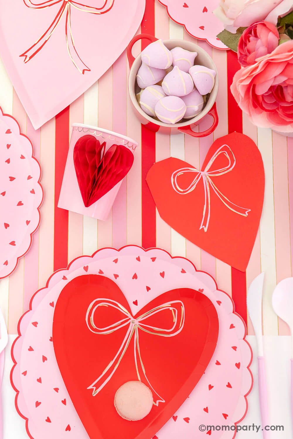 A close-up top view of Momopartys's sweet Pink and Red Bow-Themed Valentine's Day Party table. Featuring a delectable macaron on the Meri Meri Heart With Bow Plates layered with Heart Pattern Dinner Plates, Heart With Bow Napkins, Honeycomb Heart Cups, Blush and White Cutlery Set, Pink Bow Candles atop cupcakes, and sweets in the red Mini Cocotte. All elegantly arranged on a red-striped paper runner. Such a cute idea for elevating your Valentine's celebration or Galentine's Day with your besties.