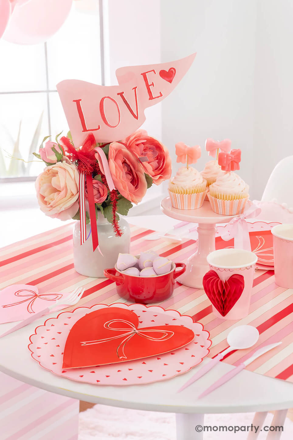 Momopartys presents A Bow-Themed Valentine's Day Table: This Bow-tastic Table Setting is filled with Meri Meri Heart Pattern Dinner Plates layered with Bow Plates and matching napkins, Honeycomb Heart Cups, Blush and White Cutlery Set, Pink Bow Candles on cupcakes, and a Love Party Pennant in the flower vase, all elegantly arranged on a red-striped paper runner. What a modern and cute idea for your Valentine's or Galentine's celebration with besties."