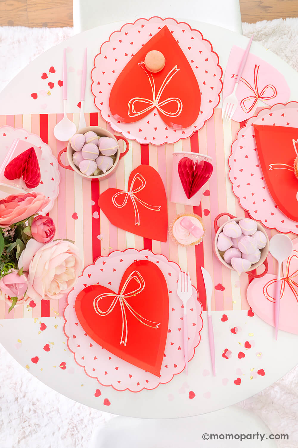 Top view of Momopartys's sweet Pink and Red Bow-Themed Valentine's Day Party table. Featuring Meri Meri Heart With Bow Plates layered with Heart Pattern Dinner Plates, Heart With Bow Napkins, Honeycomb Heart Cups, Blush and White Cutlery Set, Pink Bow Candles atop cupcakes, and sweets in the red Mini Cocotte. All elegantly arranged on a red-striped paper runner, sprinkled around Kisses Artisan Confetti. Such a cute idea for elevating your Valentine's celebration or Galentine's Day with your besties