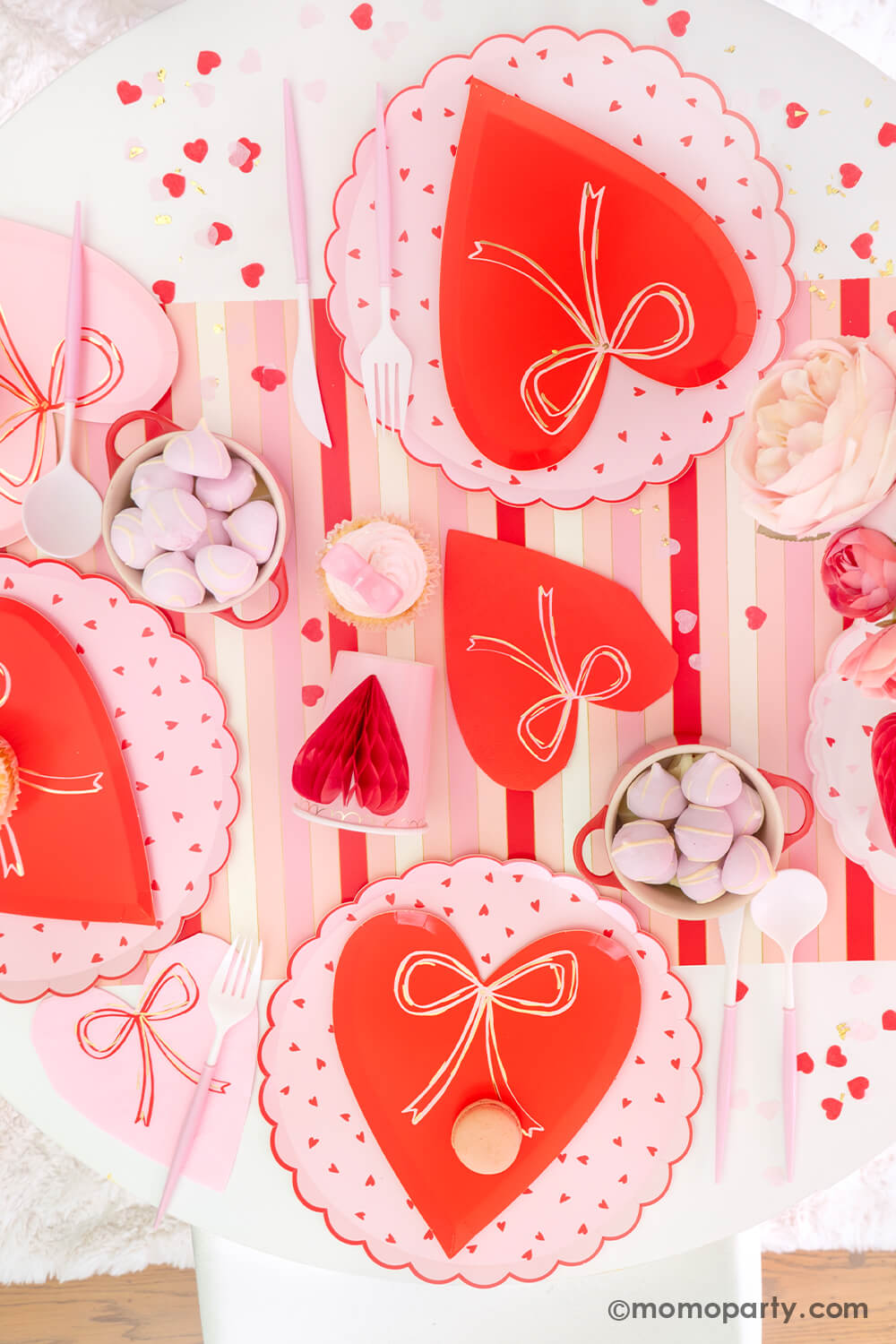 Top view of Momopartys's sweet Pink and Red Bow-Themed Valentine's Day Party table. Featuring Meri Meri Heart With Bow Plates layered with Heart Pattern Dinner Plates, Heart With Bow Napkins, Honeycomb Heart Cups, Blush and White Cutlery Set, Pink Bow Candles atop cupcakes, and sweets in the red Mini Cocotte. All elegantly arranged on a red-striped paper runner, sprinkled around Kisses Artisan Confetti. Such a cute idea for elevating your Valentine's celebration or Galentine's Day with your besties