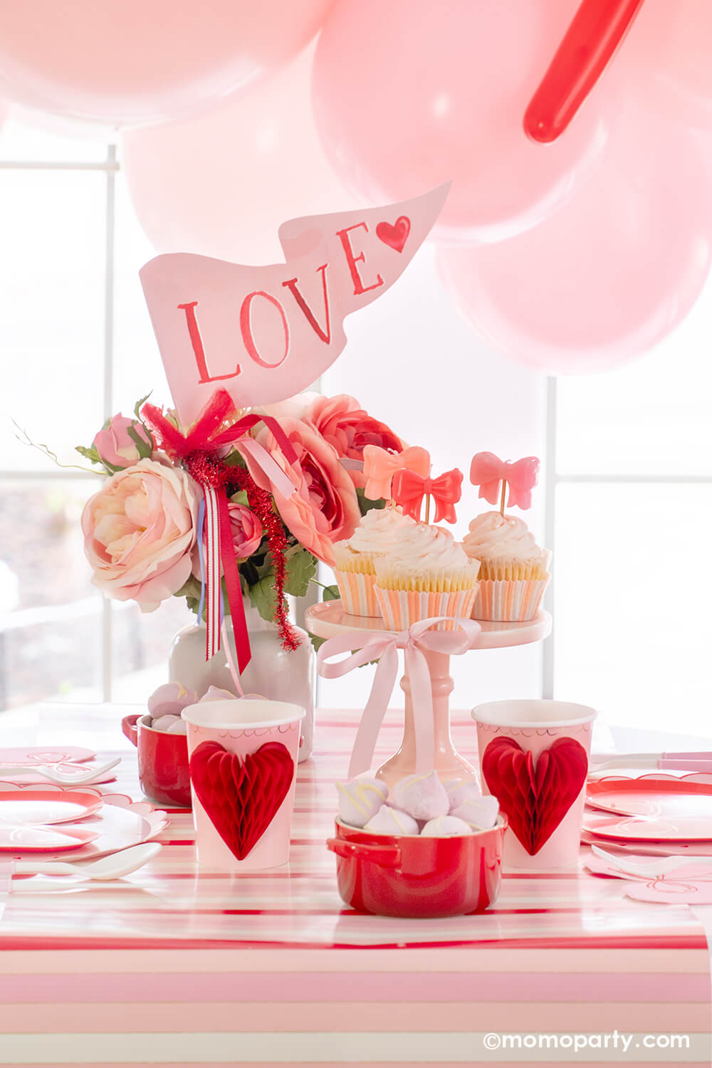 Momopartys presents A Bow-Themed Valentine's Day Party Set up. This Bow-tastic Table Setting is filled with heartwarming details, featuring Meri Meri Heart Pattern Dinner Plates layered with Bow Plates and matching napkins, Honeycomb Heart Cups, Pink Bow Candles atop cupcakes, and a Love Party Pennant adorning the flower vase, all elegantly arranged on the red-striped paper runner. It's a modern and cute idea to elevate your Valentine's celebration or Galentine's Day with your besties. 