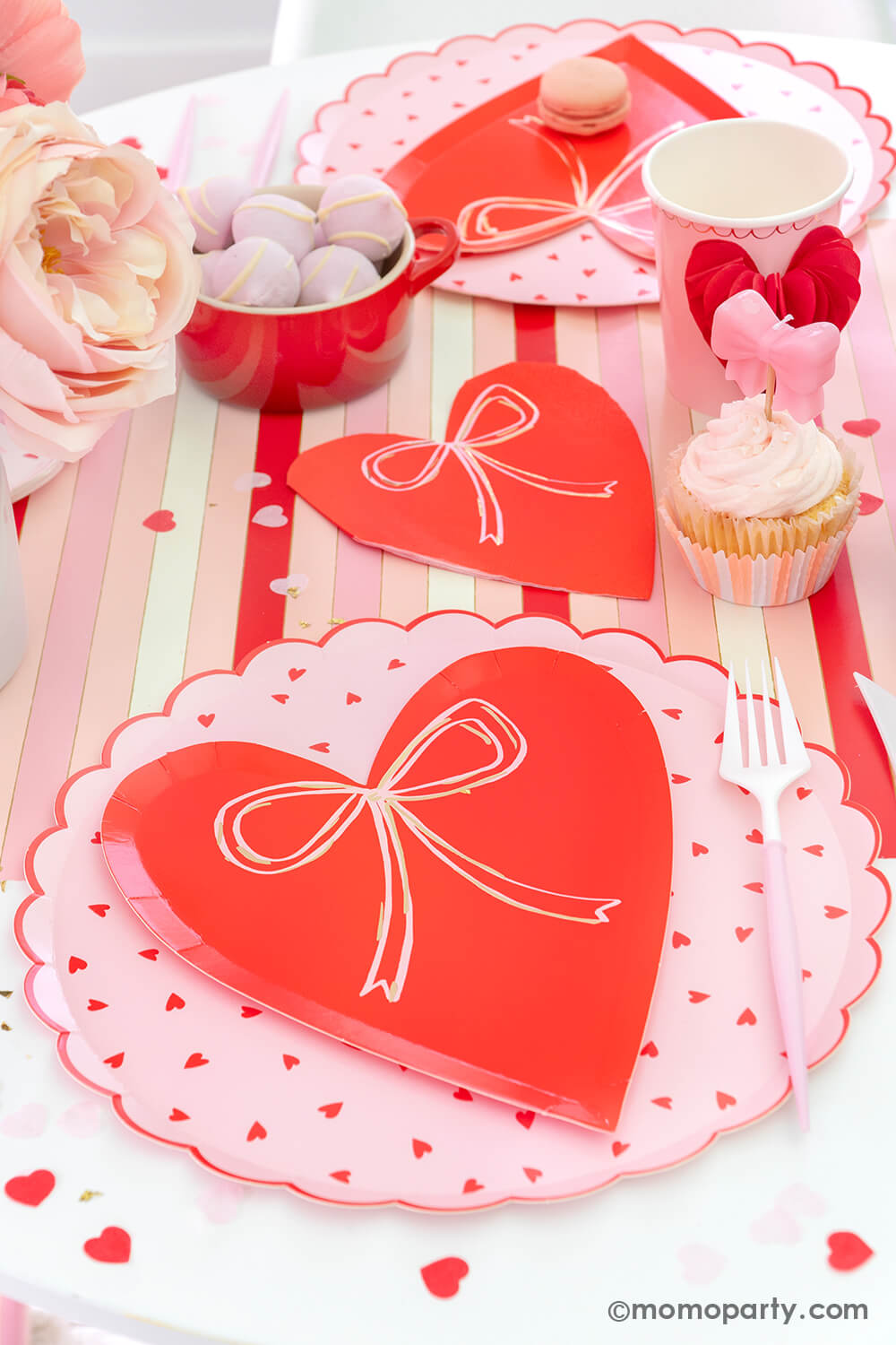 Momopartys presents a sweet Pink and Red Bow-Themed Valentine's Day Party table. Featuring Meri Meri Heart With Bow Plates layered with Heart Pattern Dinner Plates, Heart With Bow Napkins, Honeycomb Heart Cups, Blush and White Cutlery Set, Pink Bow Candles atop cupcakes, and sweets in the red Mini Cocotte. All elegantly arranged on a red-striped paper runner, sprinkled around Kisses Artisan Confetti. Such a cute idea for elevating your Valentine's celebration or Galentine's Day with your besties.