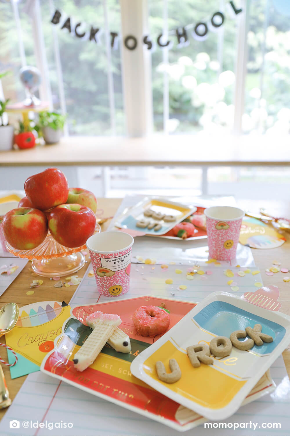 A close up shot of a festive back to school party table featuring multiple Momopartys's school themed party supplies including stack of books shaped plates, glue bottle shaped plates, crayon box shaped napkins, pink notebook party cups and giant notebook placemats. With pencil inspired confetti around and the stack of apples as the centerpiece on the table, plus the back to school banner hung above the table, this makes a perfect first day of school celebration for kids. 