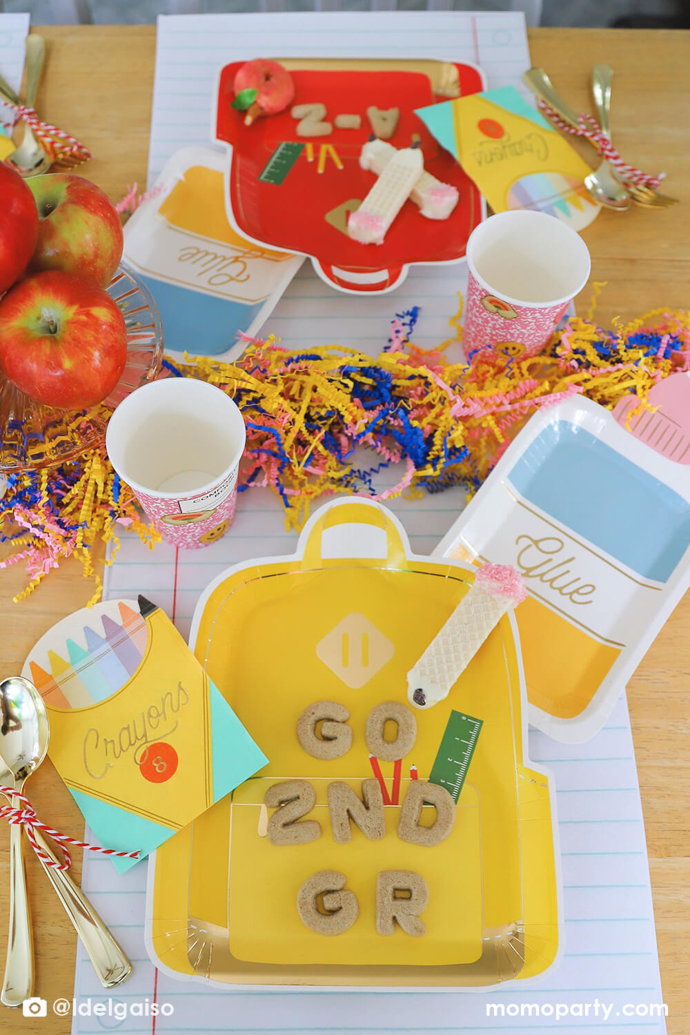 A festive back to school party tablescape featuring Momopartys's school themed party supplies including backpack shaped plate set in three different colors, glue bottle shaped plates, crayon box shaped napkins, pink notebook inspired party cups and giant notebook placemats. On the plates, there are alphabet shaped cookies spelling "Go 2nd Grader" and pencil shaped wafer cookies, with apples stacked as the centerpiece and some fun confetti, this makes a perfect first day of school celebration for kids.