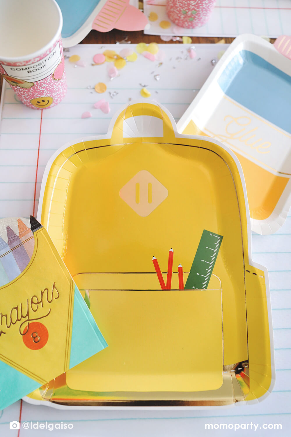 A close up shot on Momopartys's yellow backpack shaped plate by My Mind's Eye from a back to school party table featuring school themed party supplies including glue bottle shaped plates, pink notebook cups, crayon box shaped napkins and giant notebook placemats. With pencil inspired confetti, this makes a simple yet cute table setting for kid's first day of school celebration.