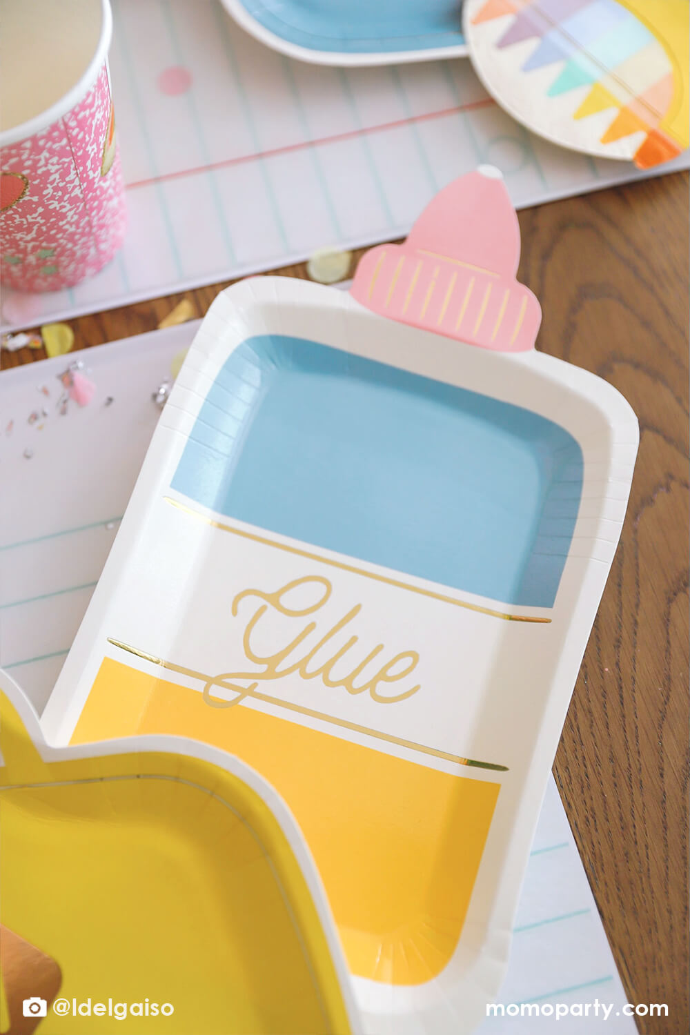 A close up shot of Momopartys's glue bottle shaped party plate by My Mind's Eye from a back to school party table. Around the glue bottle plate are some other school themed party supplies including a pink notebook party cup, a crayon box shaped napkin, the yellow backpack shaped plate on the giant notebook placemats which covered by some pencil inspired confetti.