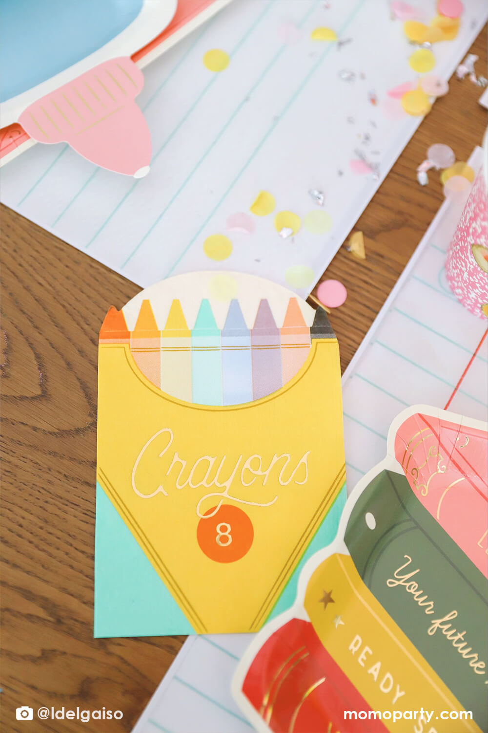 A closeup shot of Momopartys's crayon box shaped napkin next to the colorful stack of books plate on the giant notebook placemats with pencil color inspired confetti around them. These adorable school themed party supplies are essentials to your child's back to school or first day of school celebration.
