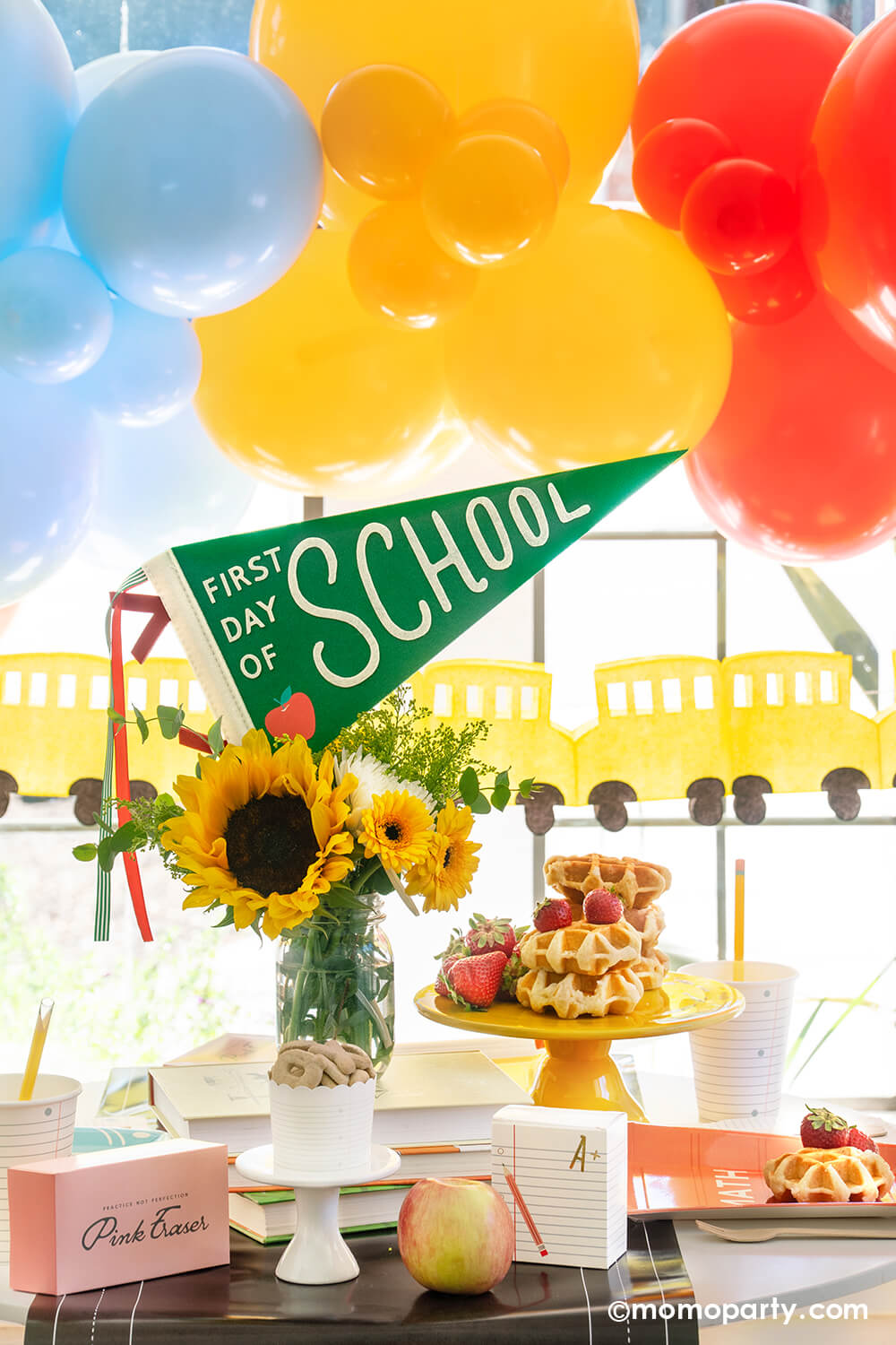 An adorable first day of school breakfast table setting featuring school themed party goods from Momopartys including the first day of school felt party pennant, chalkboard table runner, pink eraser shaped treat boxes, notebook shaped treat boxes, text book shaped plates, notebook paper cups and pencil straws. There are also some waffles stacked and strawberries on a cake stand. Behind the table is a festive balloon garland and school bus party banner, a perfect set up for your child's first day of school.