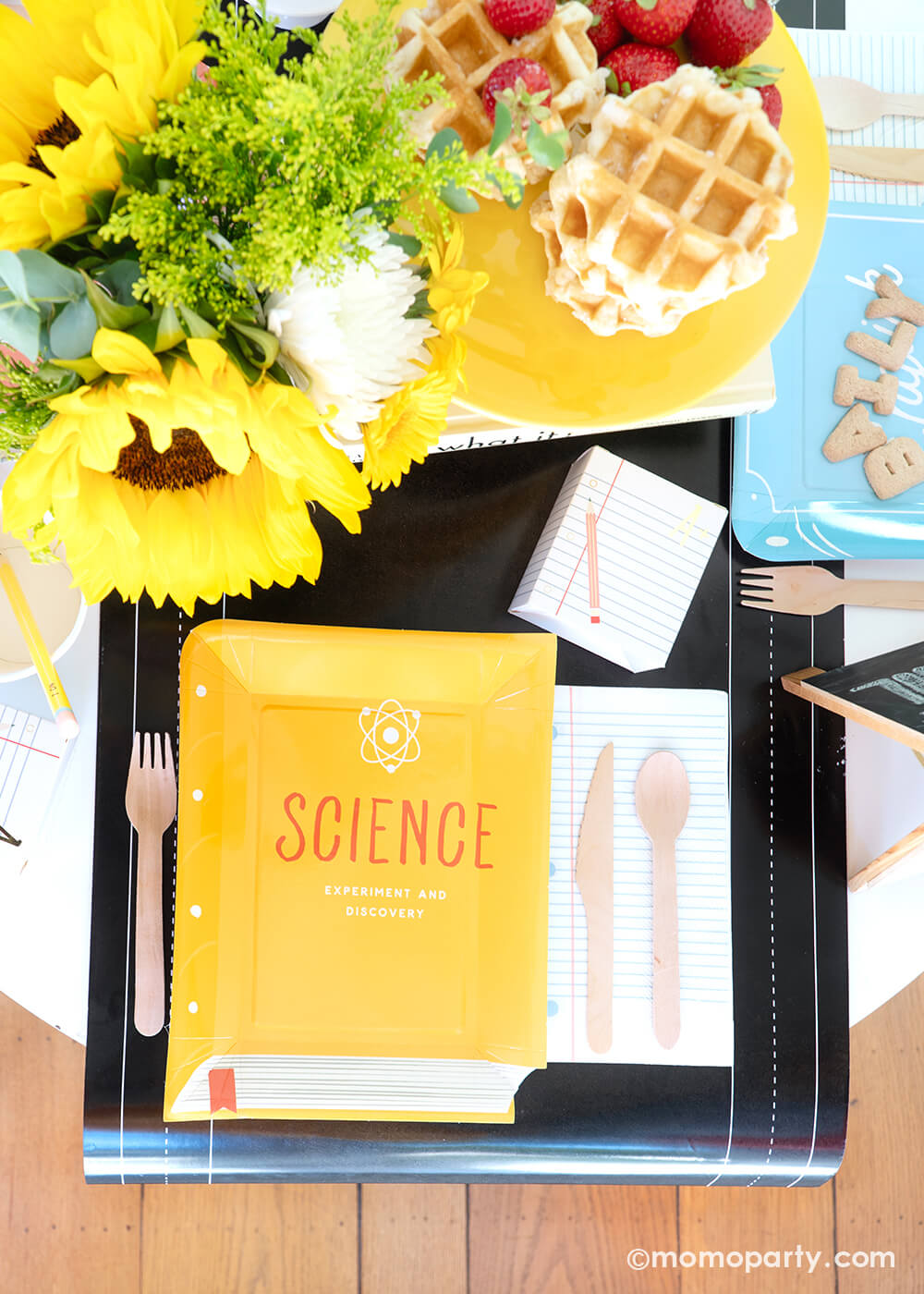 An overhead shot of a back to school party tablescape featuring multiple school themed party goods from Momopartys, including text book shaped plate in different yellow, the notebook napkin and a treat box, all on the chalkboard table runner. With stacked waffles on a cake stand, fruits, alphabet cookies, and a sunflower floral arrangement on the table, this makes a perfect idea for your child's back to school party or first day of school celebration!