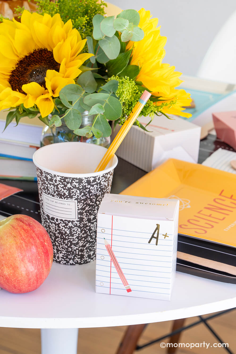 An close-up shot of a back to school party table featuring multiple school themed party goods from Momopartys, including text book shaped plates in different colors,  composition party cups with pencil straws, and ruled notebook treat boxes, all on the chalkboard table runner. With apples and a sunflower floral arrangement on the table, this makes a perfect idea for your child's back to school party or first day of school celebration!