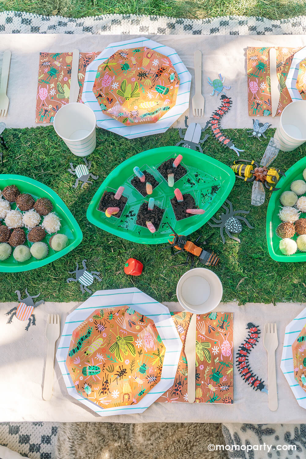 An insect bug themed birthday party table styled by Momopartys features backyard bug plates, napkins with aqua striped plates and party cups by Daydream Society. In the middle there's a faux grass table runner that was decorated with various lego insect toys and palm leaf shaped serving tray filled with insect themed treats and snacks including gummy worms, ladybug babybel cheeses and bug egg shaped brigadeiros, some playful and fun ideas for kid's insect themed bash.