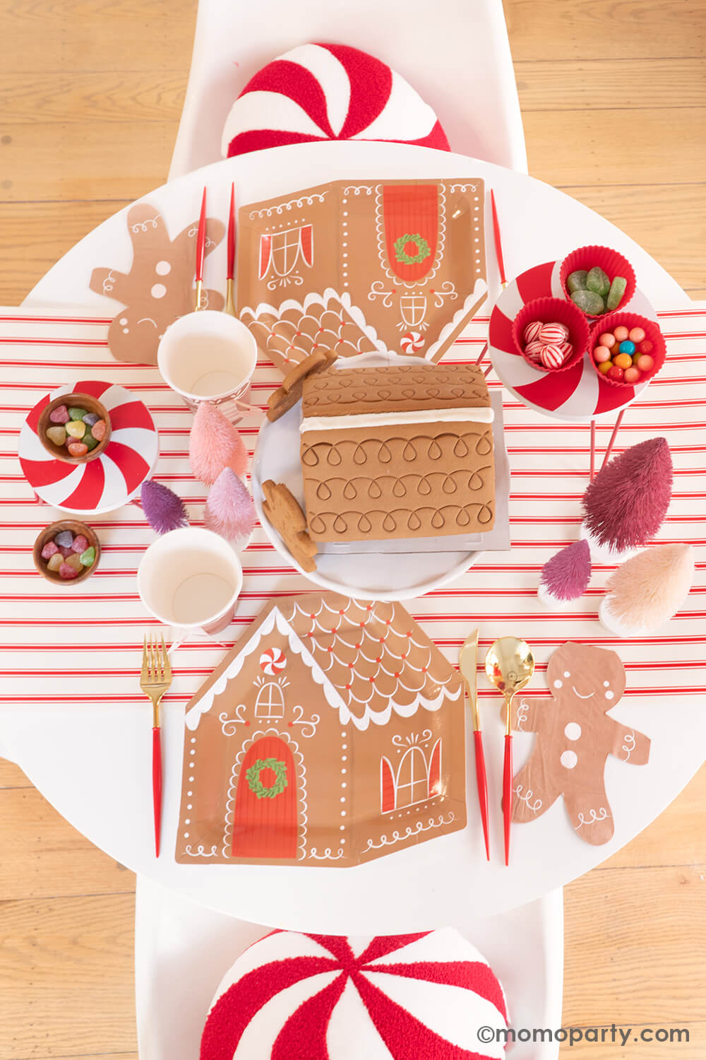 Top view of Momopartys's Gingerbread House Decoration Home Party: A dining table adorned with My Mind's Eye Gingerbread House-Shaped Plates, Smiley Gingerbread Man Napkins, Gold and Red Cutlery Sets, and Gingerbread House Paper Party Cups with Handles. A pre-built Gingerbread House sits in the center, ready for decorating, surrounded by Christmas Bottle Brush Trees and candies—all atop the festive Believe Christmas Red Striped Table Runner. What a delightful way to celebrate the holiday with kids at home!