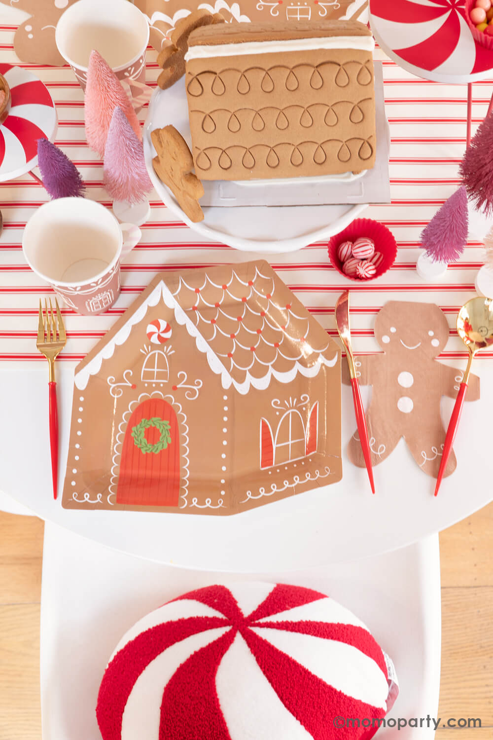 Top view of Momopartys's Gingerbread House Decoration Home Party: A dining table adorned with My Mind's Eye Gingerbread House-Shaped Plates, Smiley Gingerbread Man Napkins, Gold and Red Cutlery Sets, and Gingerbread House Paper Party Cups with Handles. A pre-built Gingerbread House sits in the center, ready for decorating, surrounded by Christmas Bottle Brush Trees and candies—all atop the festive Believe Christmas Red Striped Table Runner. What a delightful way to celebrate the holiday with kids at home!