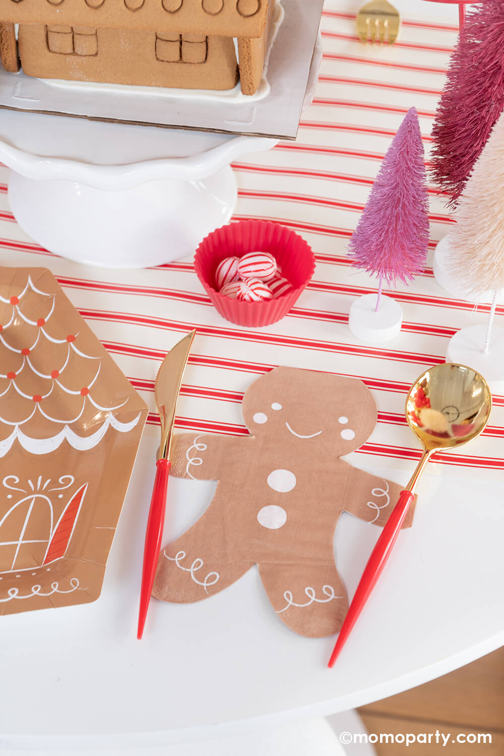 A corner of a festive holiday party table with Momopartys's smiley gingerbread man shaped napkin and festive red and gold cutlery placed on the sides on his hands next to a gingerbread house shaped plate which makes a playful tablescape. Around it is some pink bottle wash Christmas tress in different shades and some peppermint candies ready for decorating the gingerbread house for a kid's holiday fun activity. 