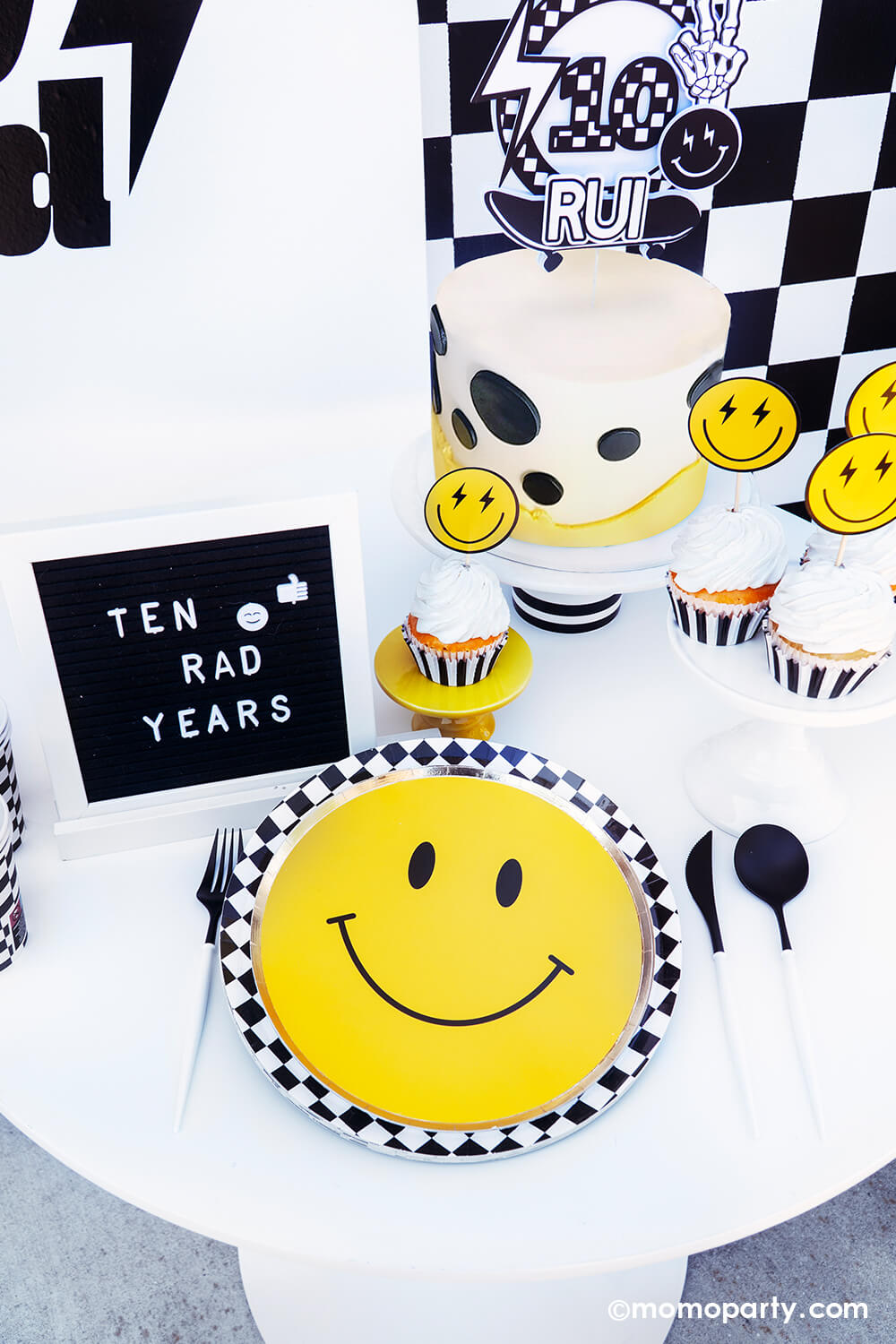 Boy's "Ten Rad Years" 10th birthday party tablescape by Momopartys. Featuring the classic black and white checkered patterned plates, cups, napkins and smiley face plates, this rad party table decoration is perfect for your cool dude's very first double digits party! With a mix of cool dude aesthetics, smiley-face accents, and checkered patterns in black and white, we've got the perfect recipe for a memorable boy's birthday bash. Don't miss out on making this party as special as your rad boy.