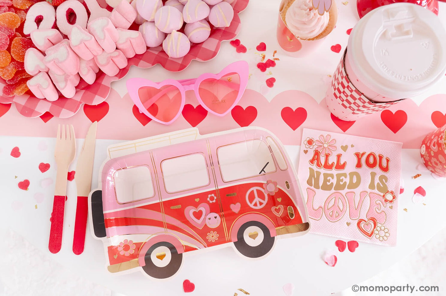 A sweet close-up of Momopartys's Groovy Valentine's Party table, showcasing My Mind's Eye's Occasions by Shakira - Luv Bus Paper Plates and All You Need is Love Napkins, complemented by a Red Wooden Cutlery Set. Next to it, a Red Checks To-Go Cup, a Pink Heart Shaped Party Sunglasses add a playful touch. At the center is a heart-shaped sweet board filled with Valentine's Day themed treats, all presented on the Pink Heart Border Paper Table Runner. A perfect inspiration for a fun Valentine's celebration!