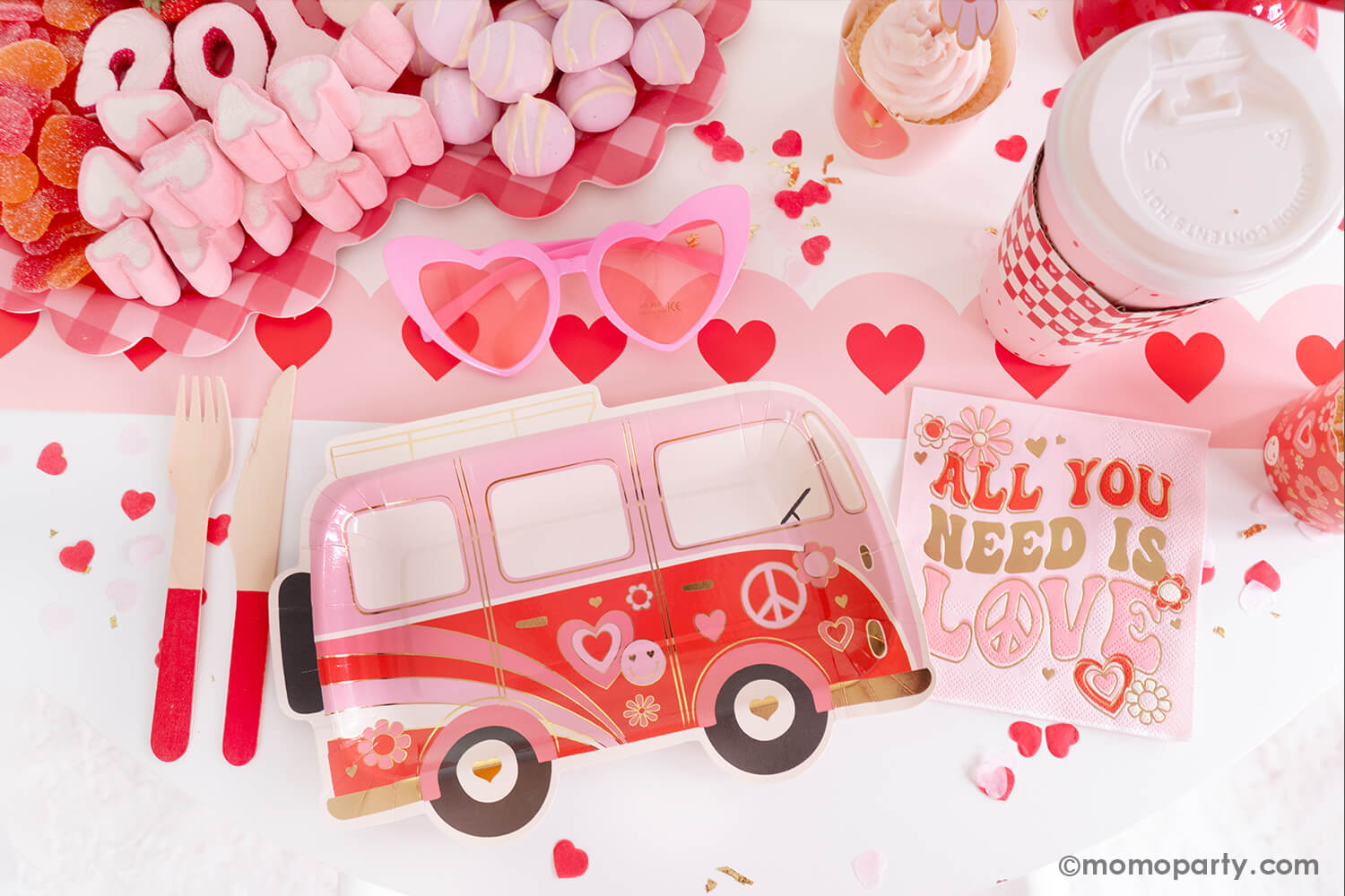A sweet close-up of Momopartys's Groovy Valentine's Party table, showcasing My Mind's Eye's Occasions by Shakira - Luv Bus Paper Plates and All You Need is Love Napkins, complemented by a Red Wooden Cutlery Set. Next to it, a Red Checks To-Go Cup, a Pink Heart Shaped Party Sunglasses add a playful touch. At the center is a heart-shaped sweet board filled with Valentine's Day themed treats, all presented on the Pink Heart Border Paper Table Runner. A perfect inspiration for a fun Valentine's celebration!