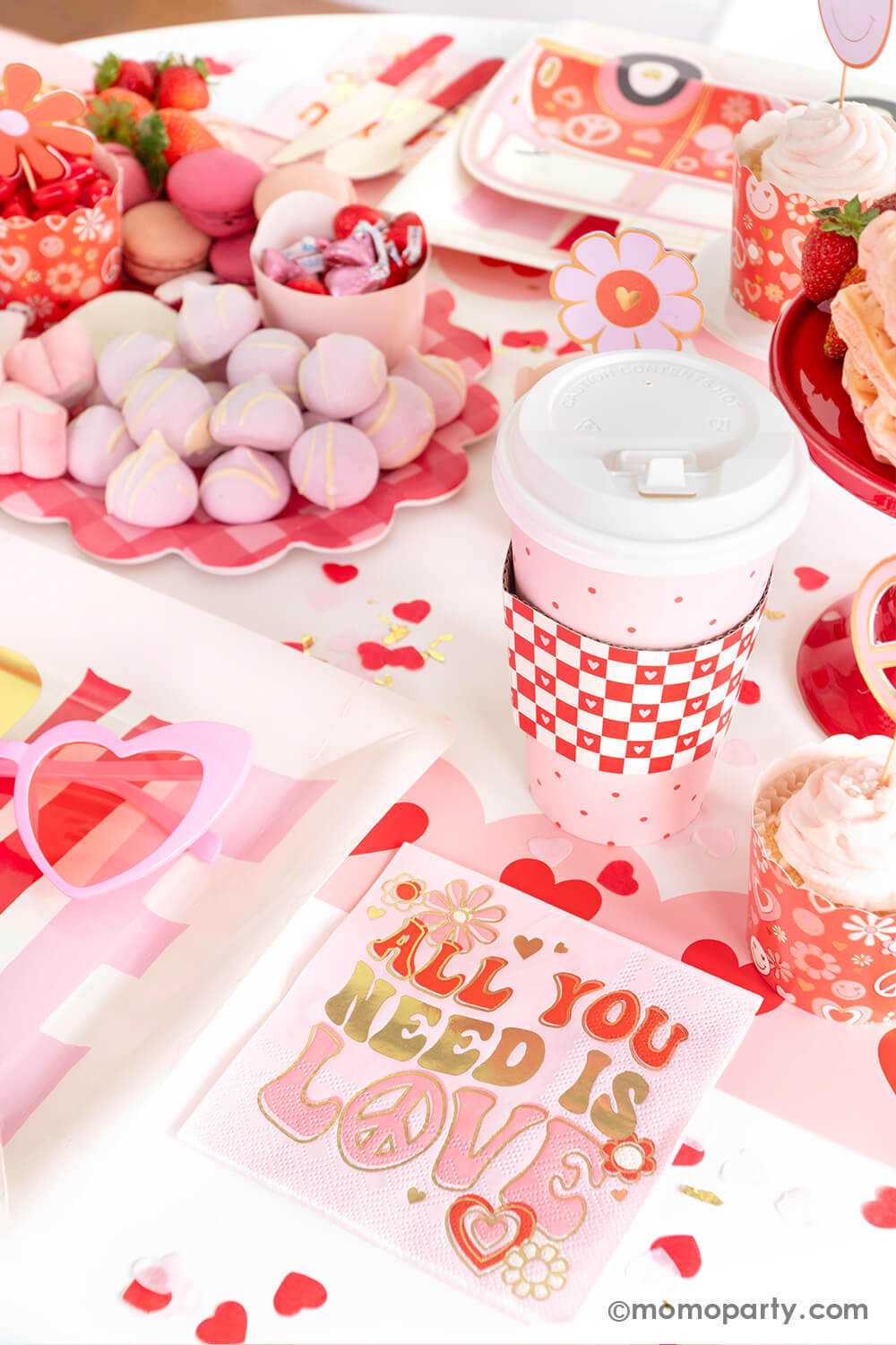 A party table featuring Momopartys's Groovy Valentine's party box with My Mind's Eye's retro Valentine's party supplies including baking cups, "all you need is love" napkins, VW bus shaped plates and red checkered party cups. On the table there's a heart shaped sweet board filled with Valentine's Day themed treats including macarons, strawberries, heart shaped marshmallows, heart shaped pink waffles and candies, a perfect inspo for a fun Valentine's celebration!