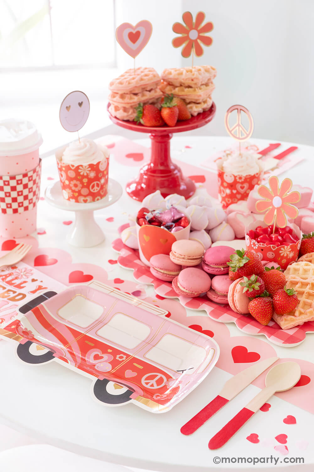 A party table featuring Momopartys's Groovy Valentine's party box with My Mind's Eye's retro Valentine's party supplies including VW bus shaped plates, red checkered party cups, baking cup and topper set and "all you need is love" napkins. On the table there's a heart shaped sweet board filled with Valentine's Day themed treats including macarons, strawberries, heart shaped marshmallows, heart shaped pink waffles and candies, a perfect inspo for a fun Valentine's celebration!