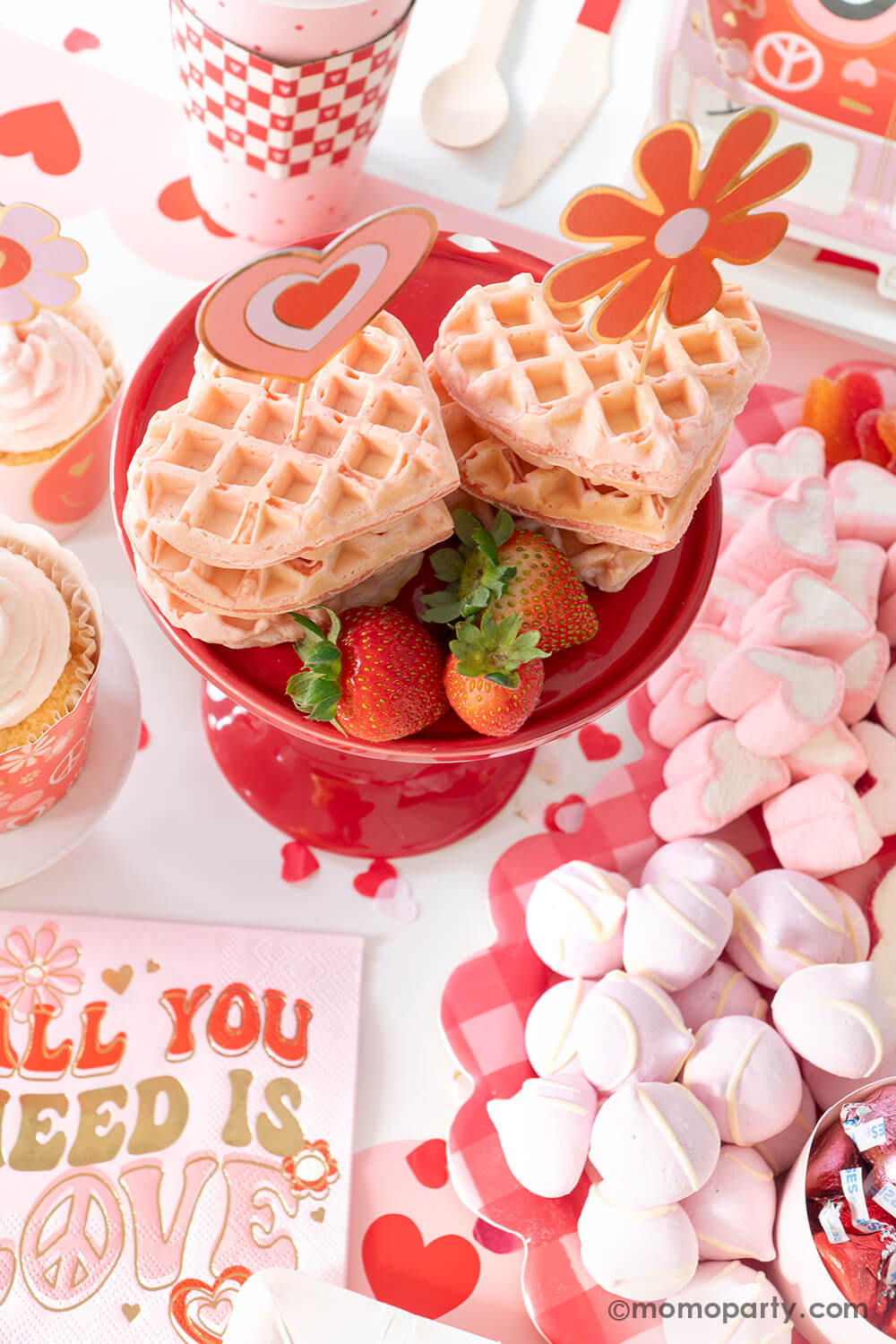 A festive Valentine's Day party table featuring a sweet board filled with heart shaped treats and desserts in pink and red. In the middle some heart shaped pink waffles topped with Momopartys's groovy love themed toppers with strawberries next to them. On the table there are lots of retro inspired Valentine's Day themed party supplies including Momopartys's all you need is love napkins, vw bus shaped plates, and red checkered party cups, makes this a perfect info for a sweet Valentine's Day celebration.