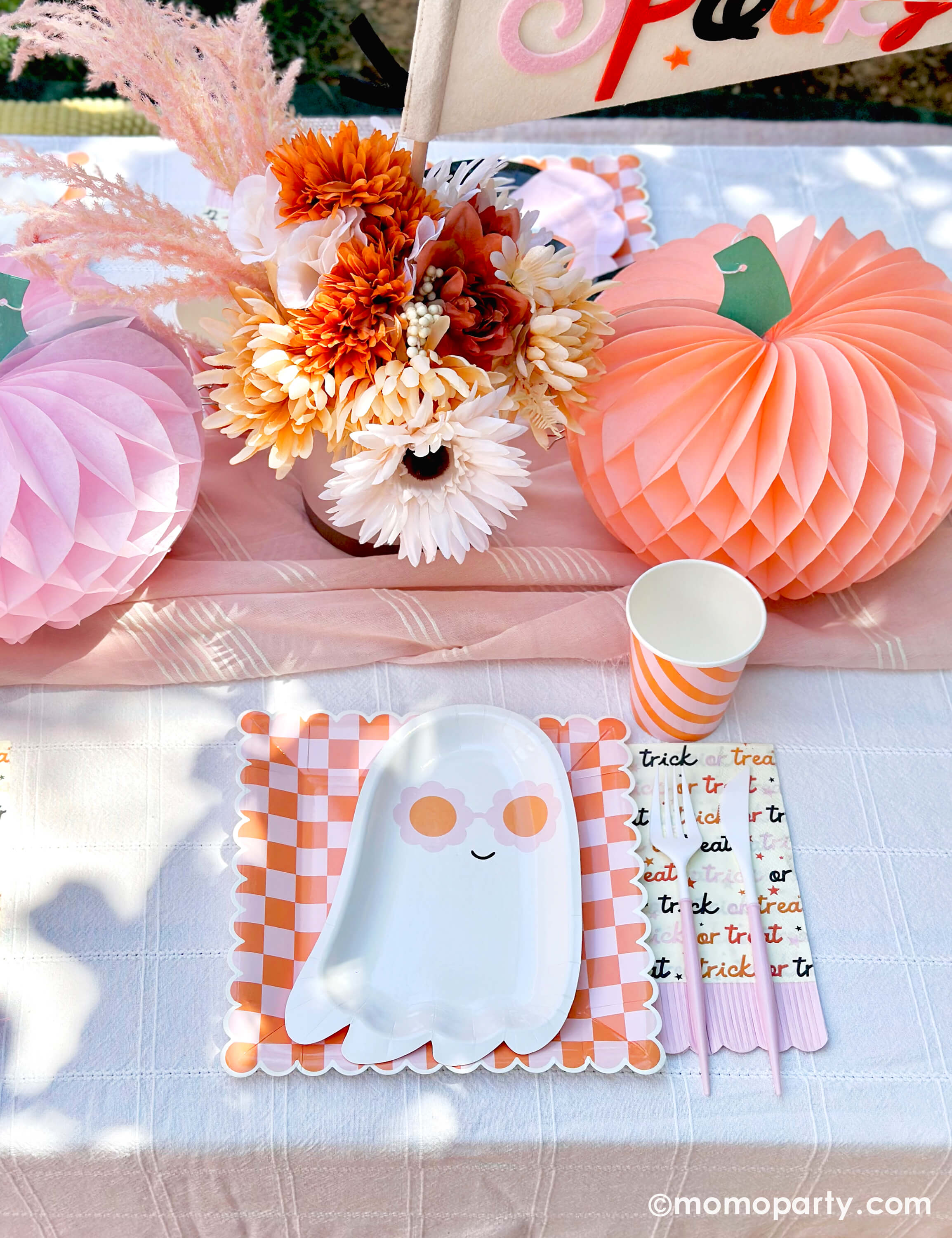 A pink Halloween table featuring Momopartys's pink sunny ghost shaped plates paired with orange/pink checkered plates, trick or treat patterned napkins with pastel pink fridge and blush and white cutlery, with boho vibe fall flower arrangement and pastel peach and pink honeycomb pumpkins as the centerpiece, and a felt party pennant that has "spooky" written on, this tablescape gives a great inspiration for a modern and chic Halloween party table decoration ideas.