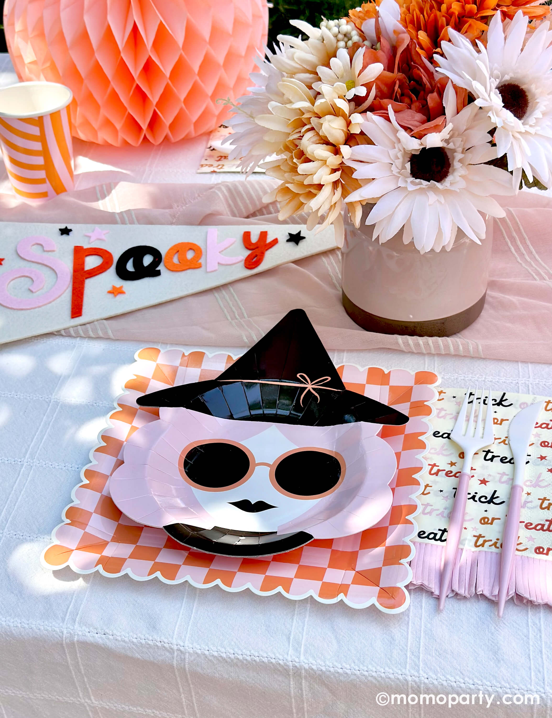 A pink Halloween table featuring Momopartys's pink witch plates paired with orange/pink checkered plates, with boho vibe fall flower arrangement and pastel peach honeycomb pumpkins as the centerpiece, and a felt party pennant that has "spooky" written on, this tablescape gives a great inspiration for a modern and chic Halloween party table decoration ideas.
