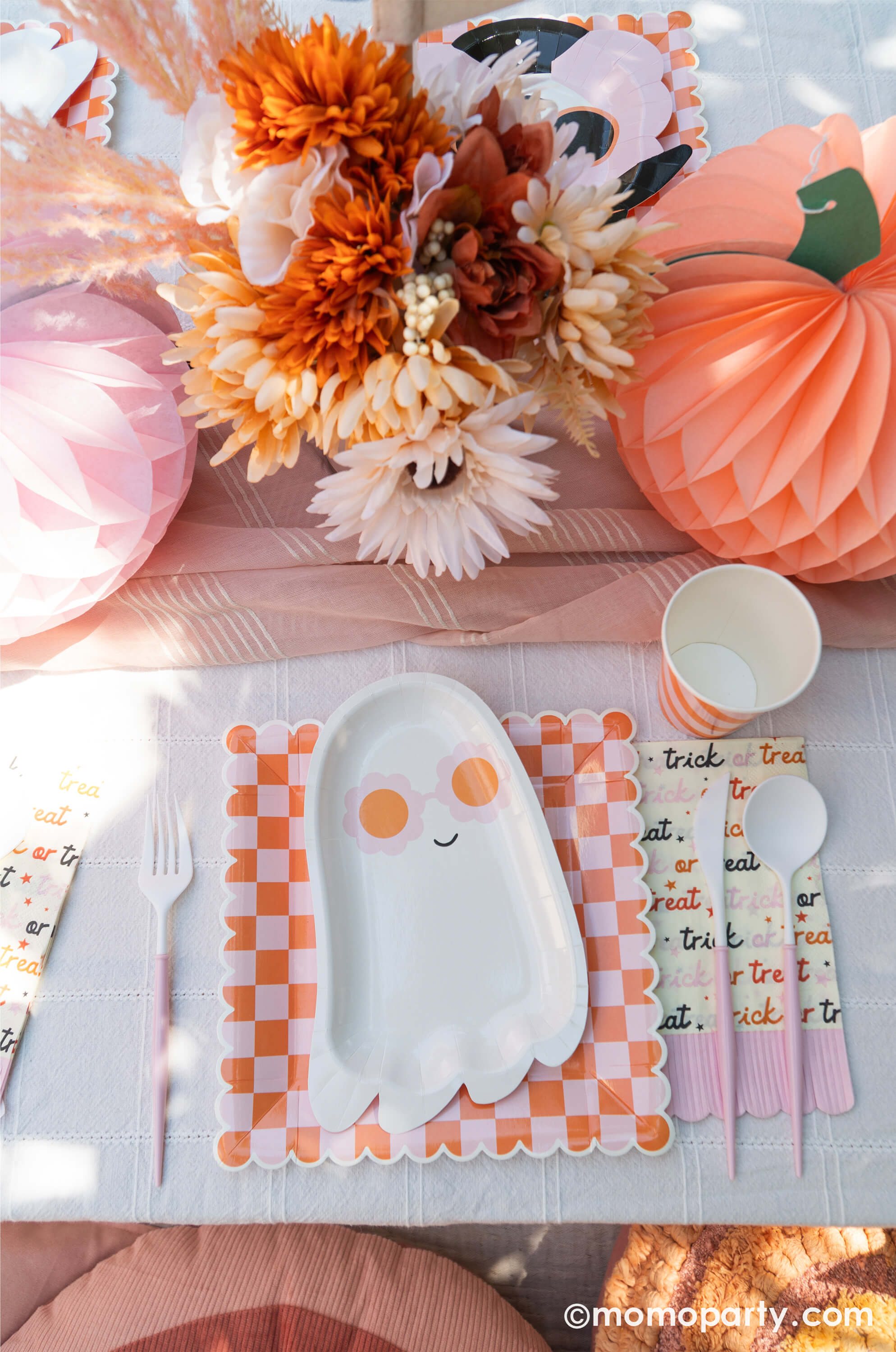 A pink Halloween table featuring Momopartys's pink sunny ghost shaped plates paired with orange/pink checkered plates, with boho vibe fall flower arrangement and pastel peach and pink honeycomb pumpkins as the centerpiece, this tablescape gives a great inspiration for a modern and chic Halloween party table decoration ideas.
