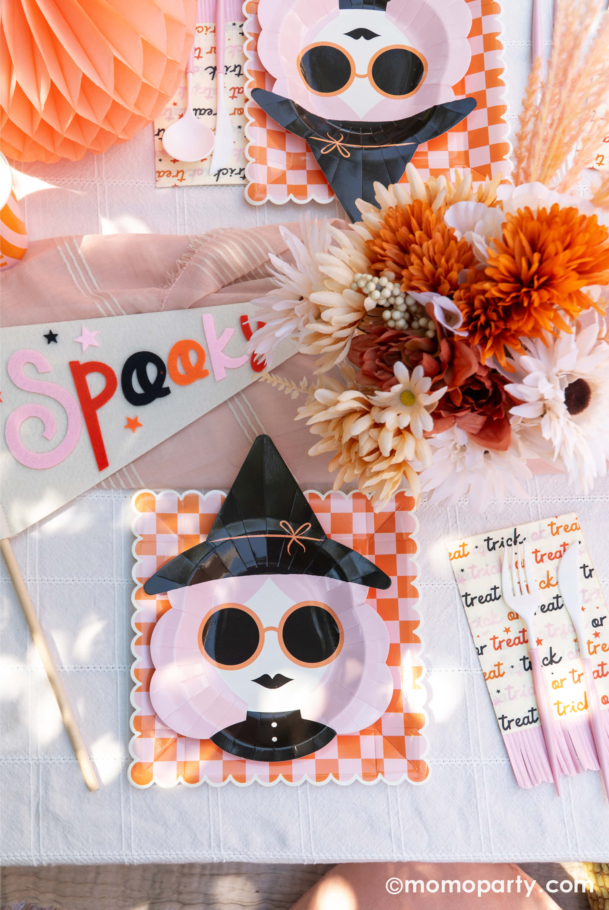 A pink Halloween table featuring Momopartys's pink witch plates paired with orange/pink checkered plates, with boho vibe fall flower arrangement and pastel peach honeycomb pumpkins as the centerpiece, and a felt party pennant that has "spooky" written on, this tablescape gives a great inspiration for a modern and chic Halloween party table decoration ideas.