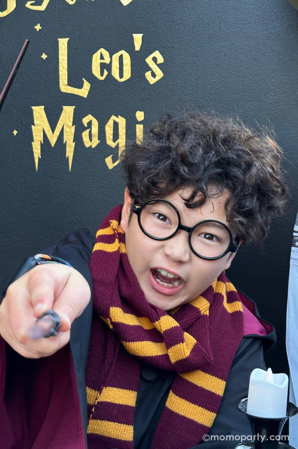 Momopartys hosted Harry Potter-themed birthday party featuring a boy dressed in a Gryffindor costume and wearing Harry Potter glasses available at momopartys.shop. Adding authentic charm to the magical theme, the boy holds a wand while casting a spell in front of a black backdrop with the text '10 years of Leo's Magic,' creating a fully immersive Harry Potter experience.