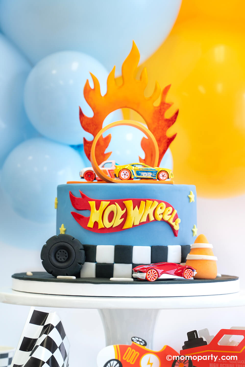 Hot Wheels themed birthday cake with the classic orange curve track and Hot Wheels toy cars by Momopartys. On the side of the cake are a fondant wheel and construction cone, making this a perfect look for kid's Hot Wheels themed birthday party.