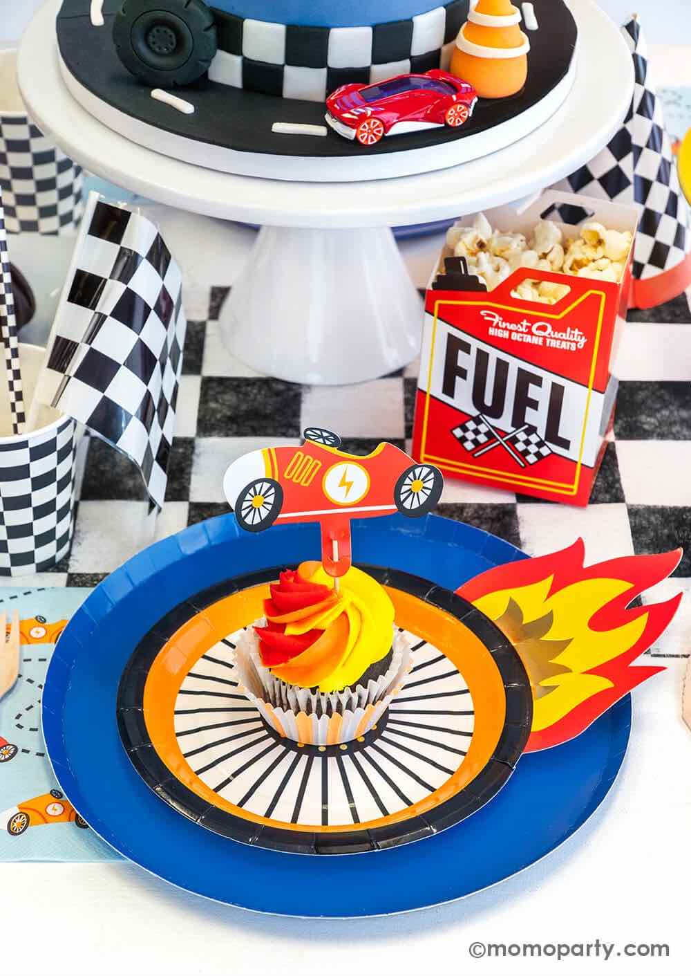 A close up shot of a Hot Wheels themed birthday party table featuring Momopartys's midnight dinner plates with wheel shaped plates, checkered table runner, party cups, paper straws and party cups, Fuel gas shaped treat box with popcorn and blue race car napkins. On the plates there's a orange and red swirl cupcake topped with vintage race car topper - a perfect inspo for boy's Hot Wheels themed birthday party.