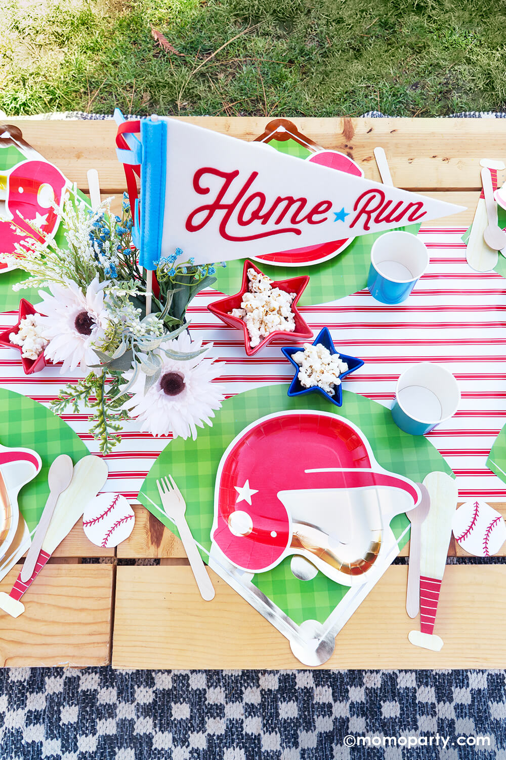 An overhead shot of kid's baseball themed birthday party picnic table setting styled by Momopartys featuring various baseball themed tablewares from Momopartys including baseball helmet shaped plate, cornflower tumbler cups, baseball shaped napkins on the baseball diamond placemats with the red pinstriped runner underneath. In the center there's a beautiful flower bouquet with the "Home Run" felt party pennant and some star shaped trays in blue and red filled with popcorn.