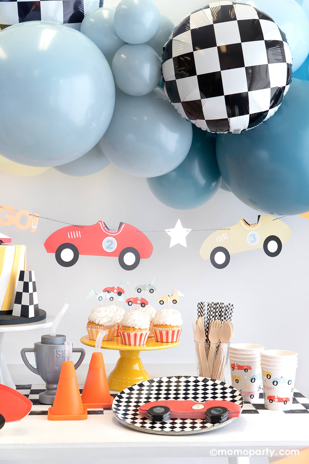 A festive race car themed party set up by Momopartys featuring a blue balloon garland adorned with checkered foil balloons, underneath hung a vintage race car party garland. On the table are some cupcakes topped with race car toppers next to a couple cone shaped and trophy shaped sippers. With checkered dinner plates, table runner and party straws, along with red race car napkins and party cups, makes a perfect inspiration for a kid's "Two Fast" second birthday party.