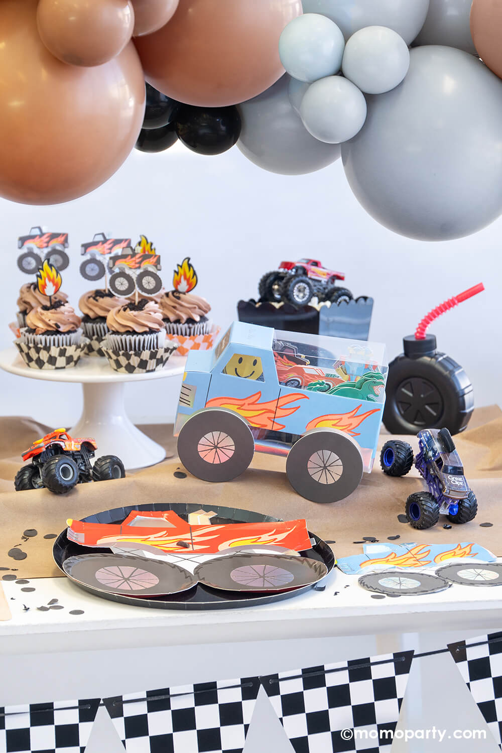 A monster truck themed party table setting styled by Momopartys decorated with party balloon cloud and checkerboard race car flags banner. The table features a red monster truck shaped paper plate with black round dinner plate, a truck shaped blue napkin, the monster truck cupcake kit and brown cupcakes topped with flame toppers, a tire shaped tumbler cup with red straw on a brown craft table cover decorated with monster truck toys and black confetti - a fun setup for kid's birthday party.
