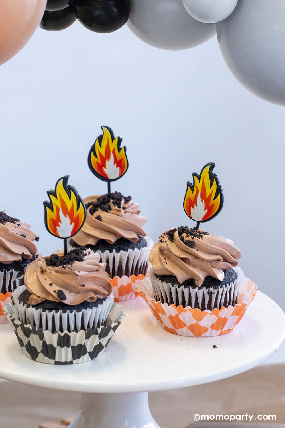 A close-up shot of a monster truck themed party decorated with party balloon garland by Momopartys. The table features a few cupcakes wrapped with orange and black checker patterned wraps with brown icing and oreo crumbs on them, each of them is topped with a fire flame topper. Making these cupcakes a fun treat idea for kid's monster truck themed birthday celebrations!