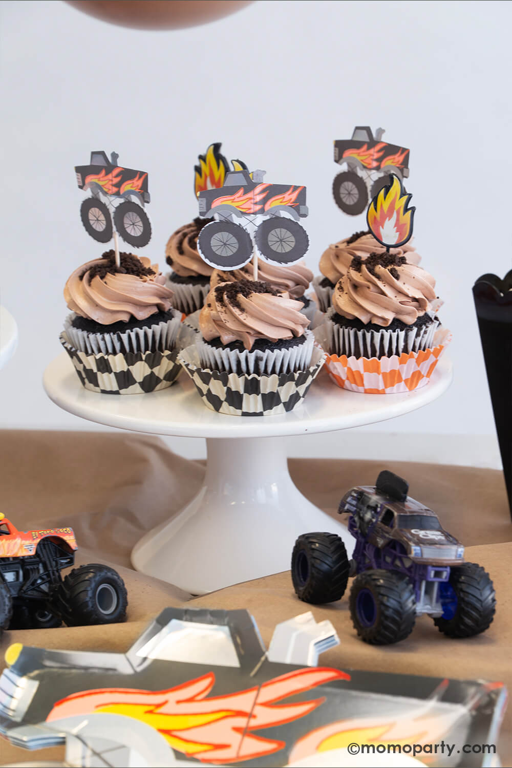 A monster truck themed party table setting styled by Momopartys. The table features a black monster truck shaped paper plate and a few cupcakes with brown icing and oreo crumbs on them, each of them is topped with either a fire flame topper or a black monster truck shaped topper. Next to the cupcakes there's a black popcorn box filled with popcorn with a monster truck toy on it. The table is covered with brown kraft paper with a few colorful monster truck toys on it, fun for kid's birthday celebrations!
