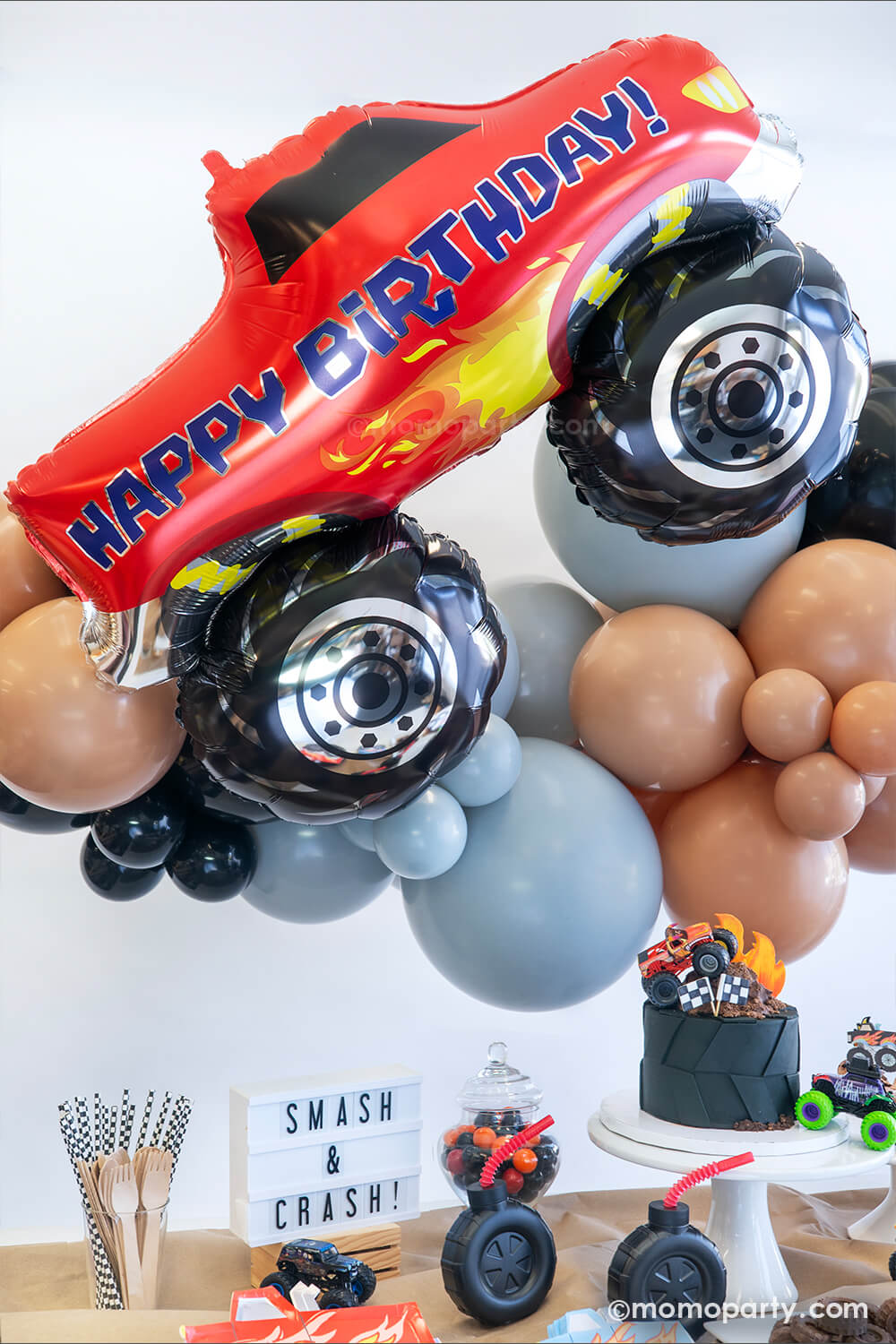 A close up of the monster truck dirt road themed balloon garland featuring various sizes of latex balloons in brown, black and grey colors with the red happy birthday monster truck shaped foil balloon by Momopartys. Underneath the balloon garland is a monster truck themed party table with a large tire shaped birthday cake and a "Smash & Crash" sign, ready for a fun and high-energy filled celebration for the little monster truck lovers!