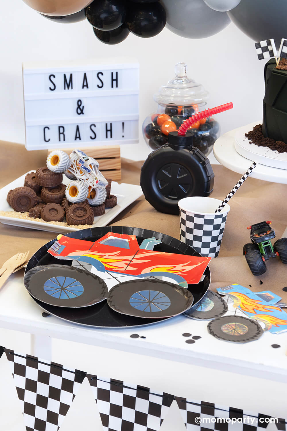 A monster truck themed party table setting styled by Momopartys decorated with party balloon cloud and checkerboard race car flags banner. The table features a red monster truck shaped paper plate with black round dinner plate, a truck shaped blue napkin, a checkerboard party cup and matching paper straw, a tire shaped tumbler cup with red straw on a brown craft table cover decorated with monster truck toys and black confetti. In the back there's a "smash & crash" sign, a fun setup for kid's birthday party.