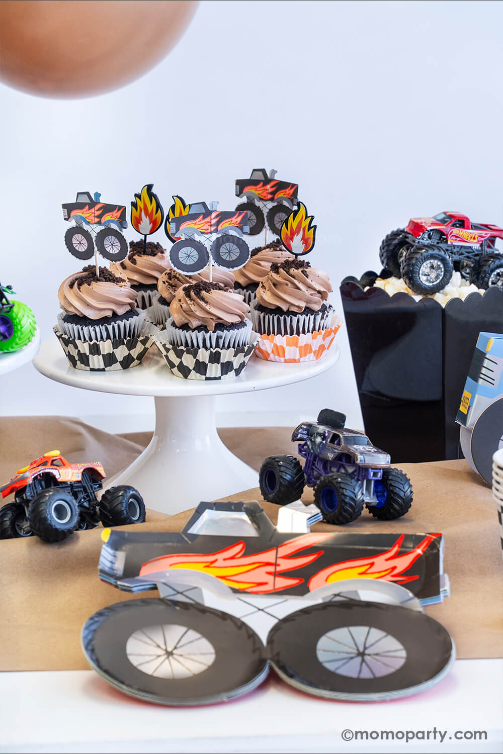 A monster truck themed party table setting styled by Momopartys. The table features a black monster truck shaped paper plate and a few cupcakes with brown icing and oreo crumbs on them, each of them is topped with either a fire flame topper or a black monster truck shaped topper. Next to the cupcakes there's a black popcorn box filled with popcorn with a monster truck toy on it. The table is covered with brown kraft paper with a few colorful monster truck toys on it, fun for kid's birthday celebrations!