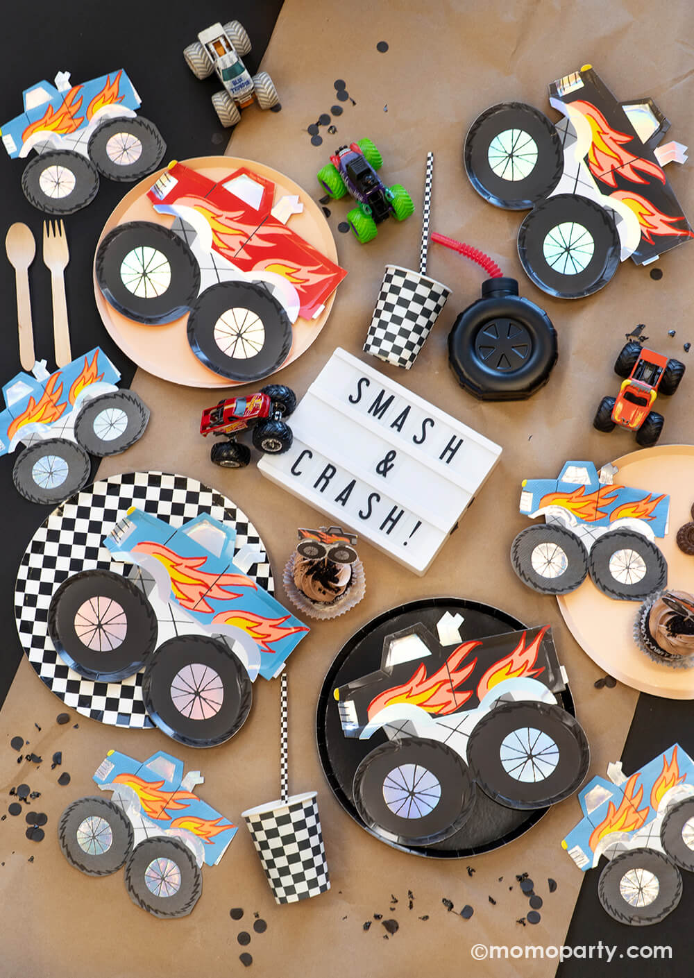 A overhead shot of a monster truck themed party tabescape featuring various monster truck themed tableware from Momopartys including round black and checkered dinner plates, party cups, straws and monster truck shaped plates and napkins, with a brown cupcake topped with a monster truck topper and "Smash & Crash" sign plus truck toys on a brown craft paper runner, this setup is perfect for your little monster truck fan's birthday celebration!