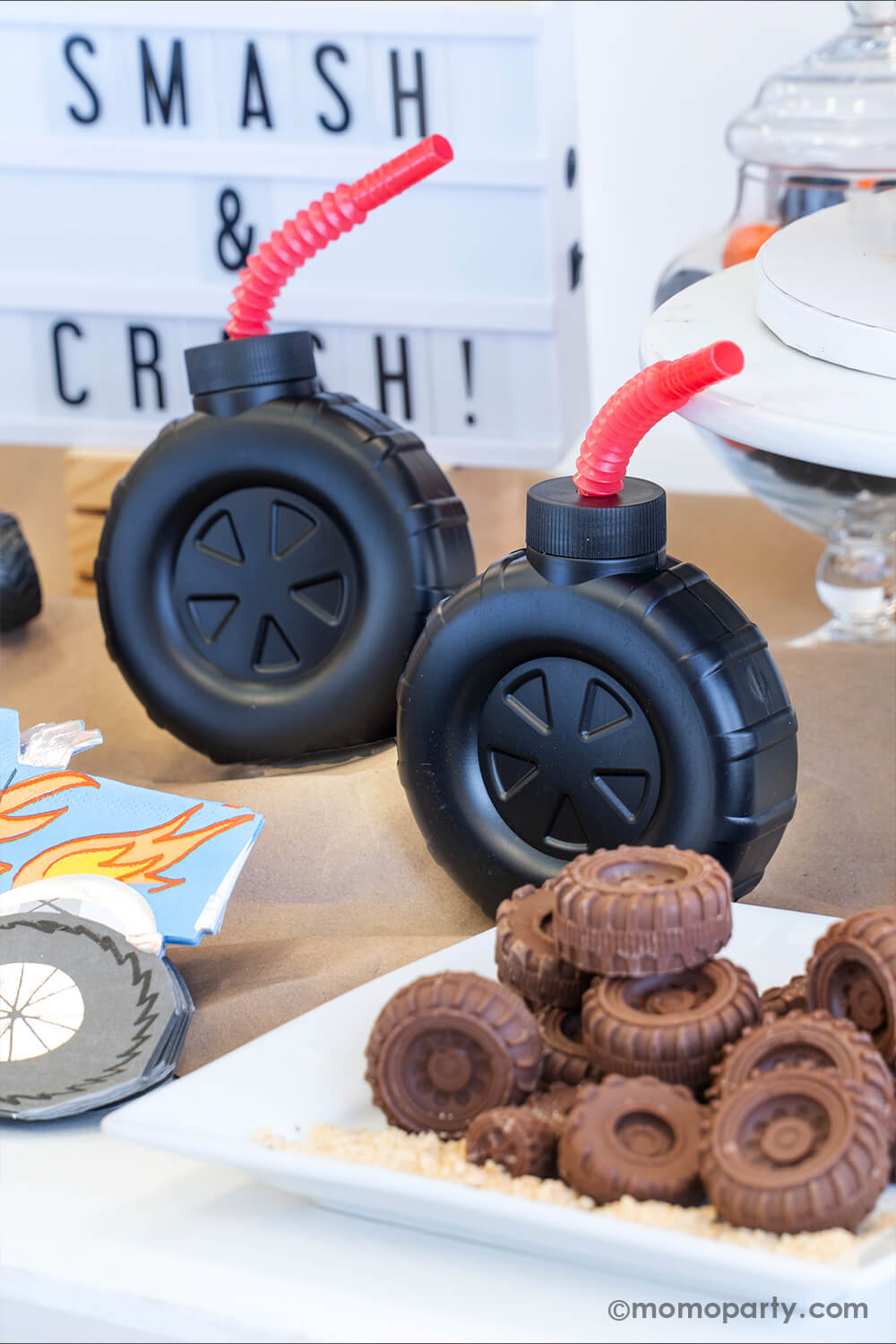 A monster truck themed party table setting styled by Momopartys. In the front there's a plate full off tire shaped chocolate candies and some graham cracker crumbs. The table is covered with brown kraft paper a couple of tire shaped tumbler cups with red straws. In the back there's a sign with "Smash & Crash" - setting up a perfect scene for a fun monster truck themed party for kids.
