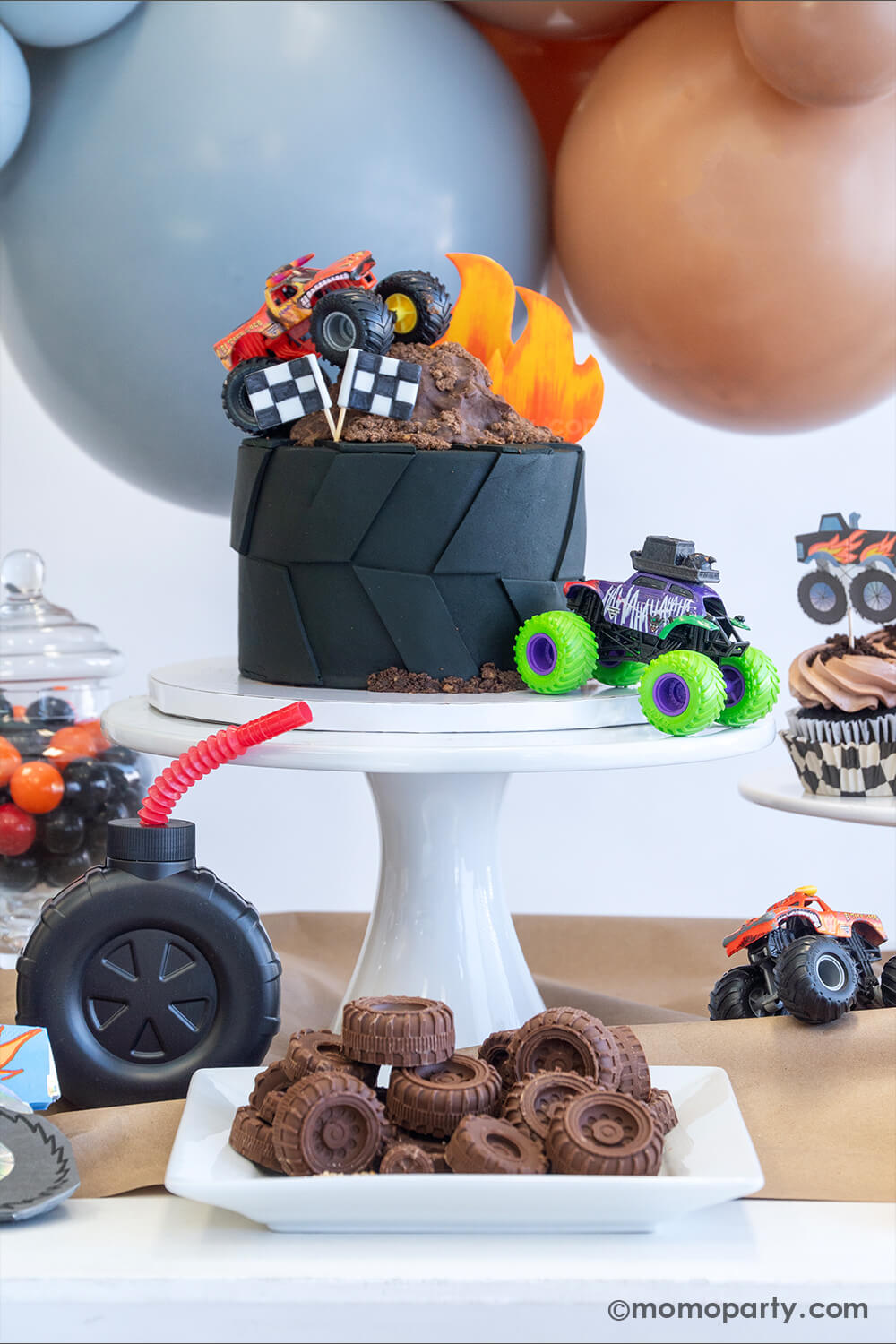 A monster truck themed party table setting styled by Momopartys. The table features a large fondant tire shaped birthday cake topped with a fondant flame, chocolate mud, two race car flags and two colorful monster truck toys. In the front there's a plate full off tire shaped chocolate candies inspired by monster truck tires. The table is covered with brown kraft paper with a candy jar with black and orange gum balls, a tire shaped tumbler cup with a red straw and a few colorful monster truck toys on it, fun