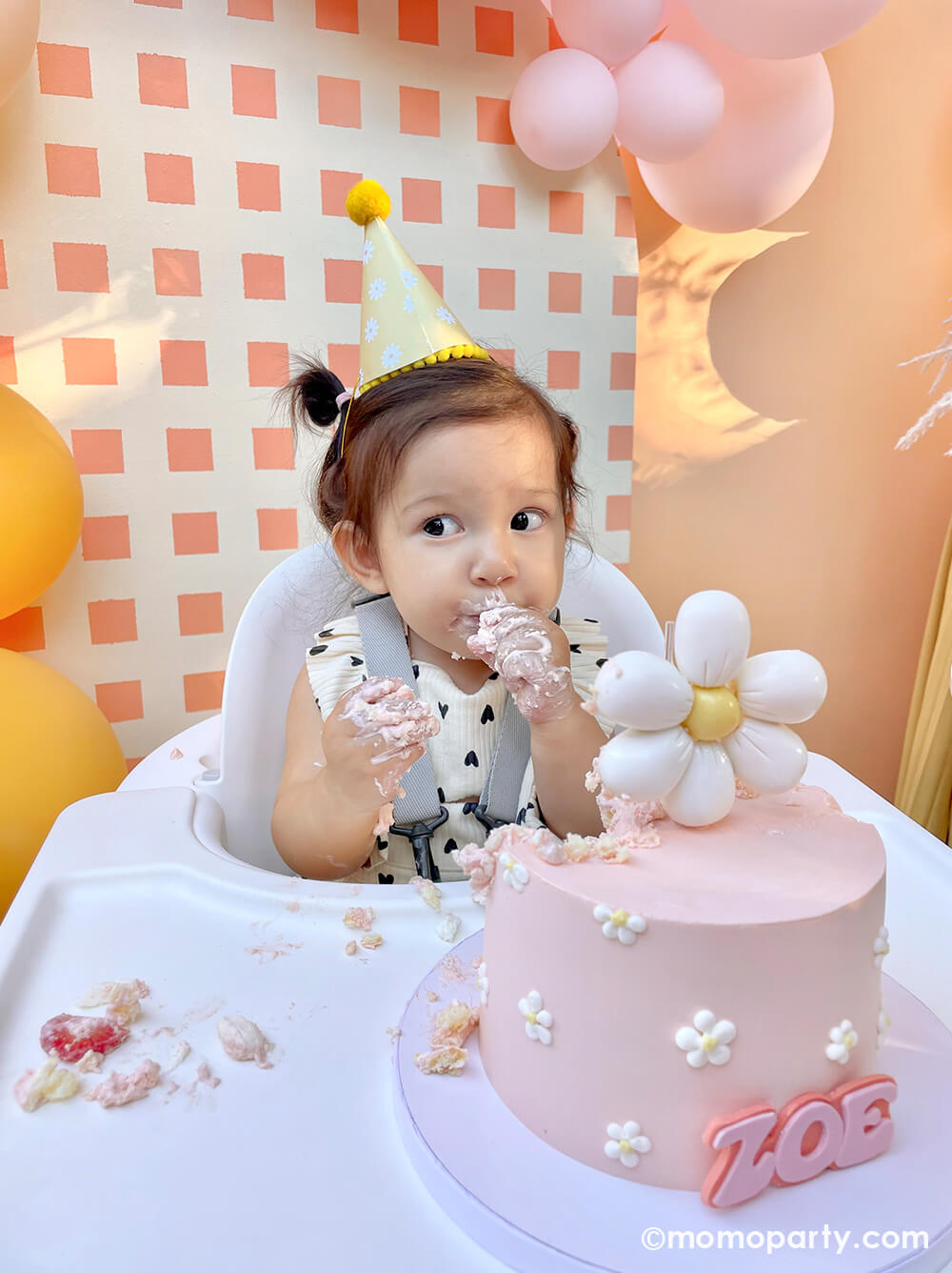 Momopartys One Groovy Baby First Birthday Party, with a birthday girl wearing a Retro Daisy Party Hat, sitting in the hair chair, eating her modern groovy-themed light pink smash cake decorated with Dasiy flower topper and Zoe in the front. She sitting in front of a check-patterned wooden Backdrop board decorated with latex balloons in peach, pink, and Goldenrod colors. This decoration is such a stand-out backdrop and photo booth for a Groovy birthday party, baby showers, and almost any other joyful event