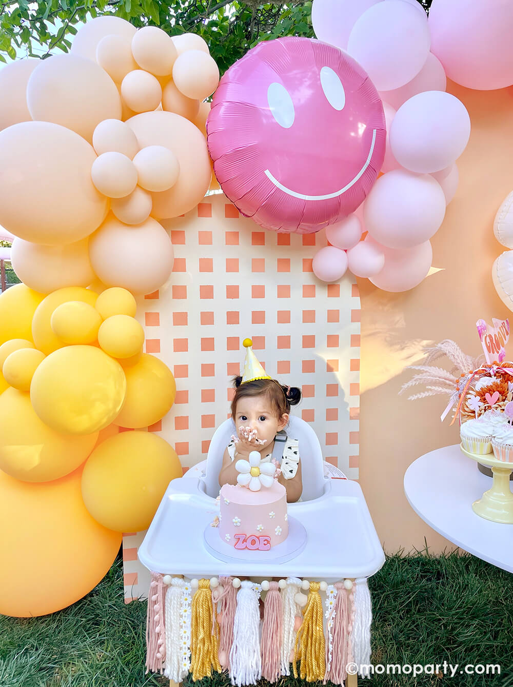 Momopartys One Groovy Baby First Birthday Party, with a birthday girl wearing a Retro Daisy Party Hat, sitting in the hair chair, eating her smash cake with a check patterned wooden Backdrop board, 30" Rosy Smiley face foil balloon by Tuftex Balloons, latex balloon decorations in peach, pink, Goldenrod colors. This decoration is such a stand-out backdrop and photo booth for a Groovy birthday party, baby showers, and almost any other joyful event