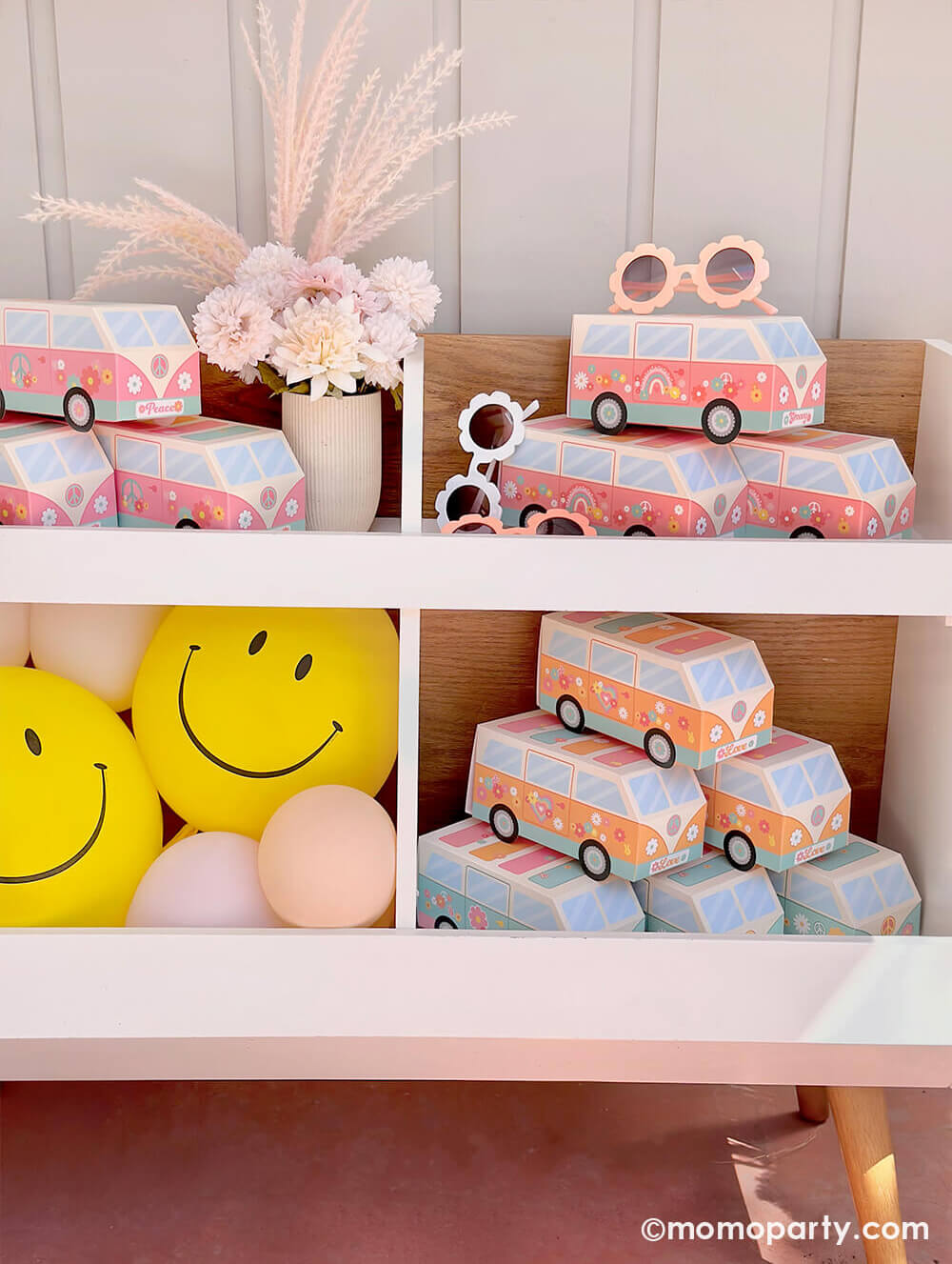 A book shelf filled with hippie themed VW van shaped treat boxes in pink and orange, along with Momopartys's yellow smiley face latex balloons at a baby girl's "Groovy One" themed first birthday party.
