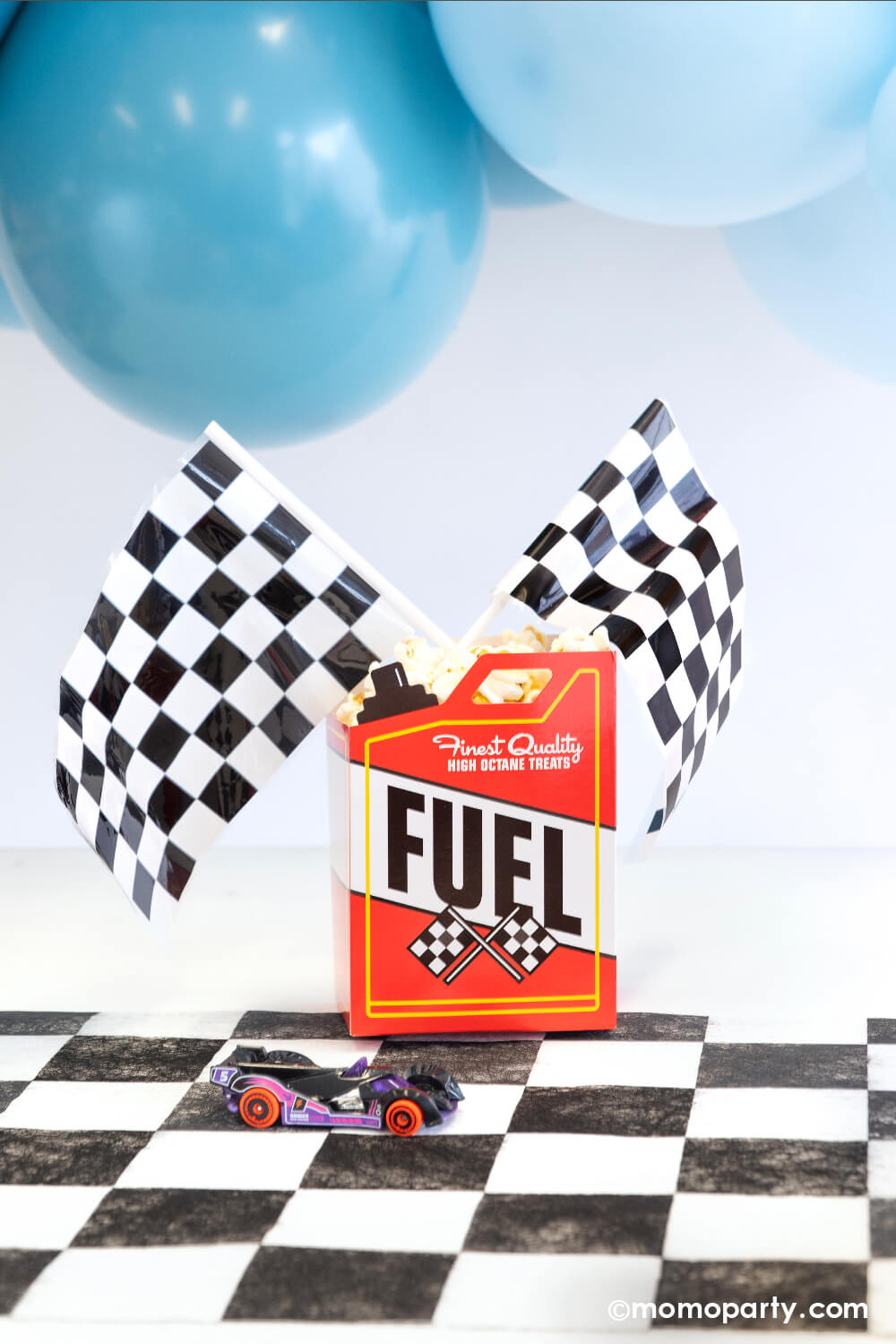 A fuel-treat-box topped with race car checkered flags from Momopartys with popcorn in side. It's setting on a checkered paper table runner with blue balloons hung on top - making this a perfect snack ideas for kid's race car themed birthday or a Hot Whees themed bash.